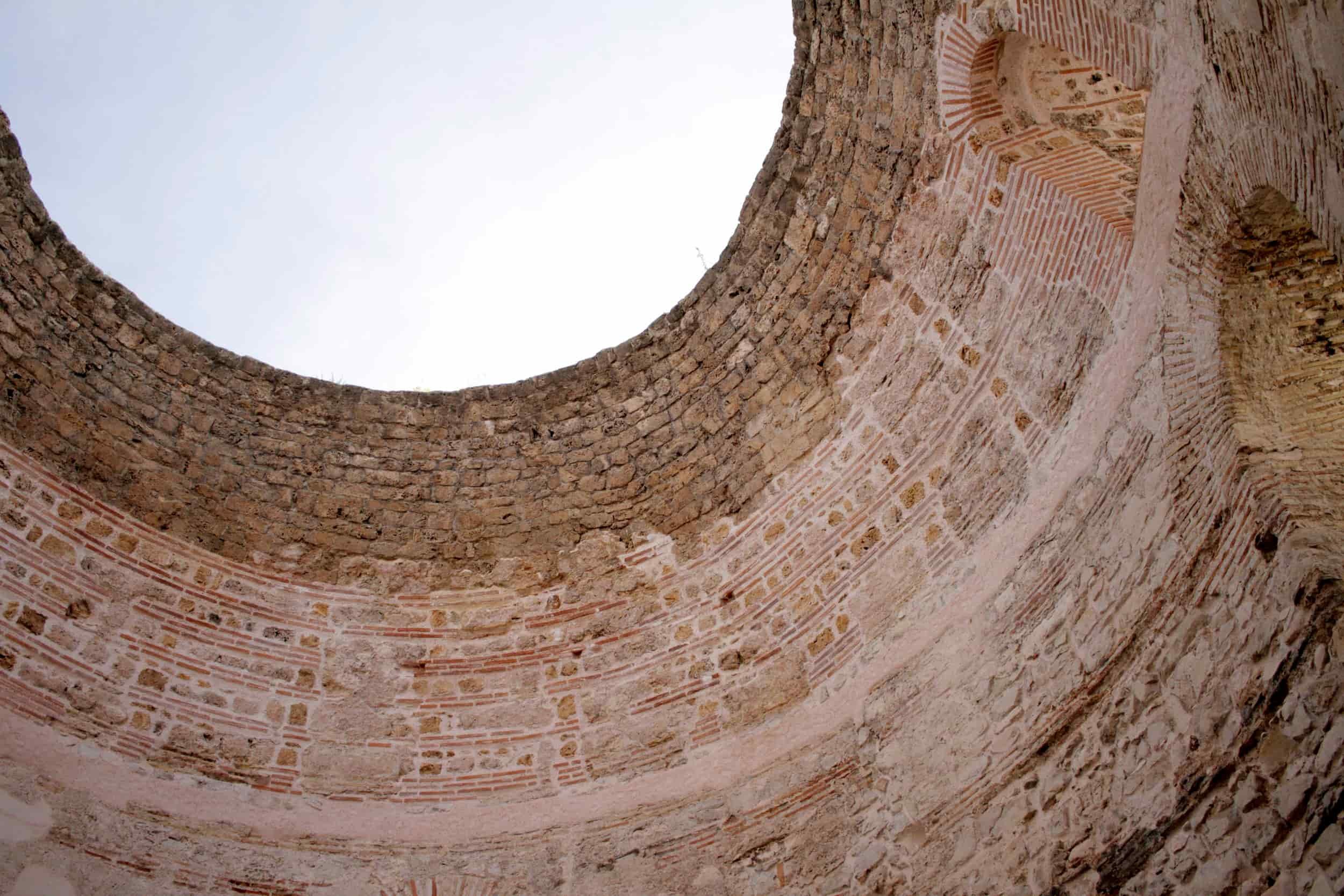 Looking up at the interior of an ancient brick and stone circular structure with an open roof, showing weathered brick and stone walls and a clear sky above.