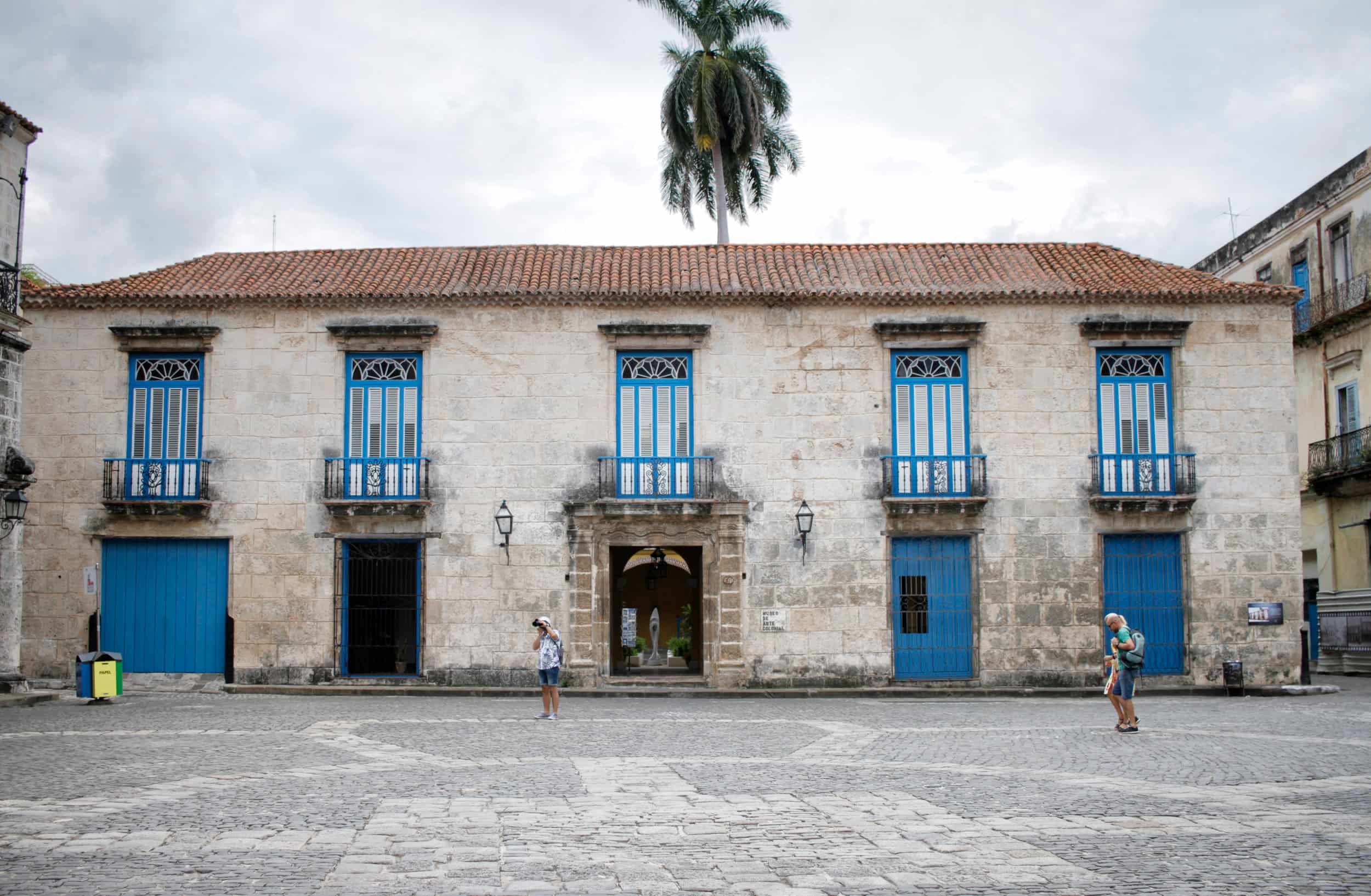 A historic stone building with tall blue windows and doors, a red tile roof, and a central arched entrance, located in a cobblestone plaza with two people standing nearby, and a tall palm tree behind the building under a cloudy sky. Havana, Cuba.