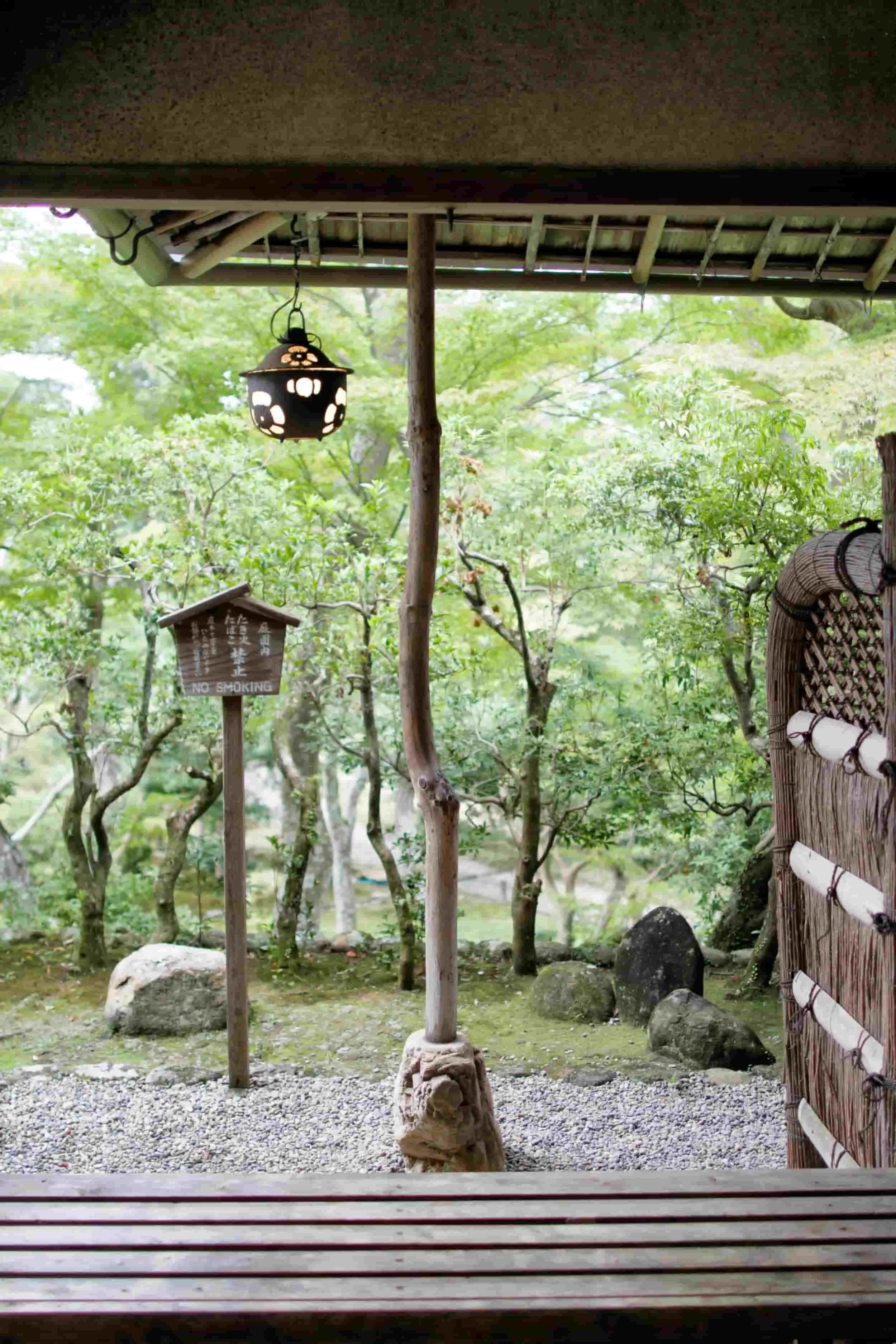 View from a shaded porch looking out into a green garden with trees, rocks, and a traditional Japanese lantern hanging from the roof.