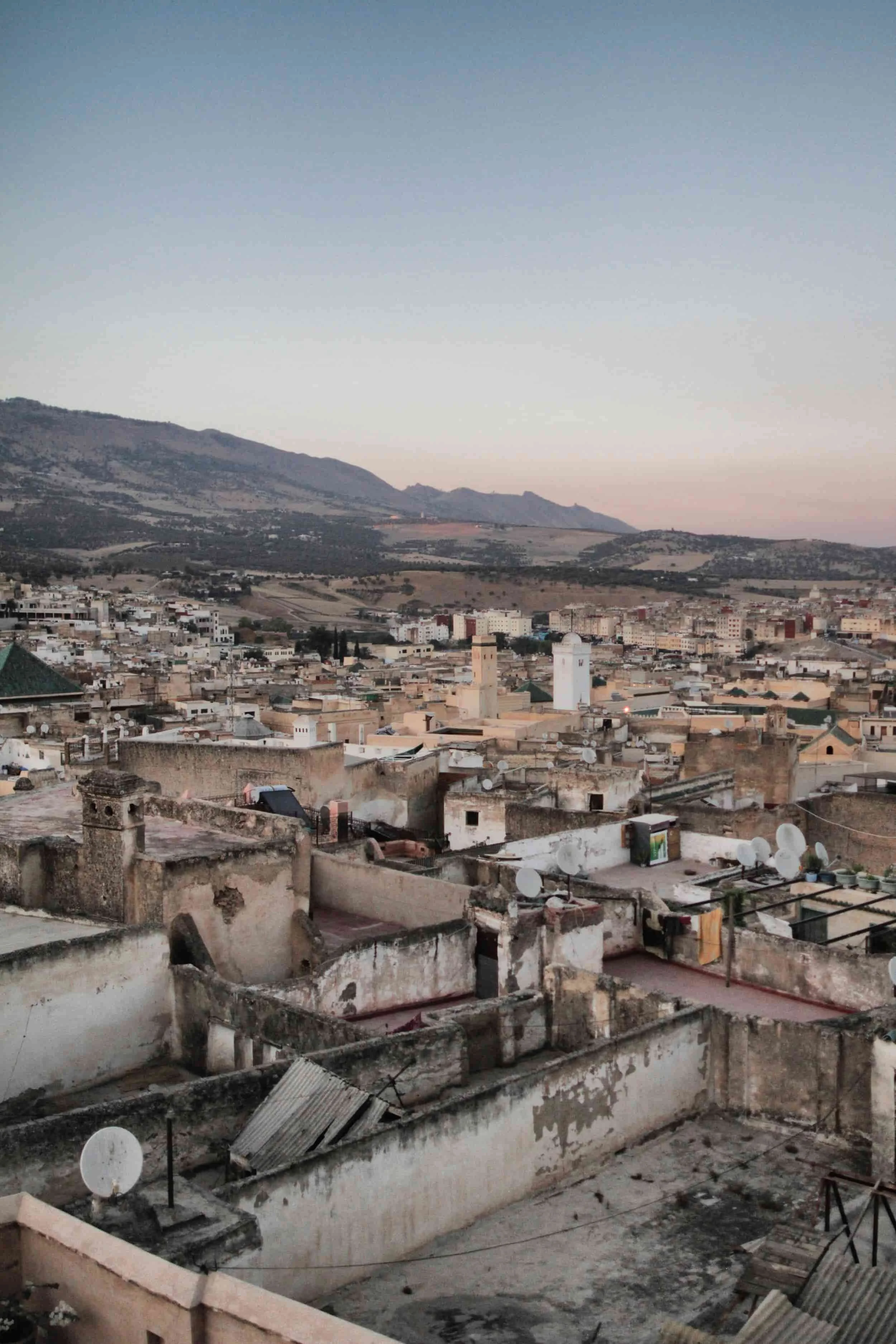 A cityscape with a mix of old and new buildings, rooftops with satellite dishes, and a mountainous landscape in the background during dusk. Fes el Bail in Morocco.