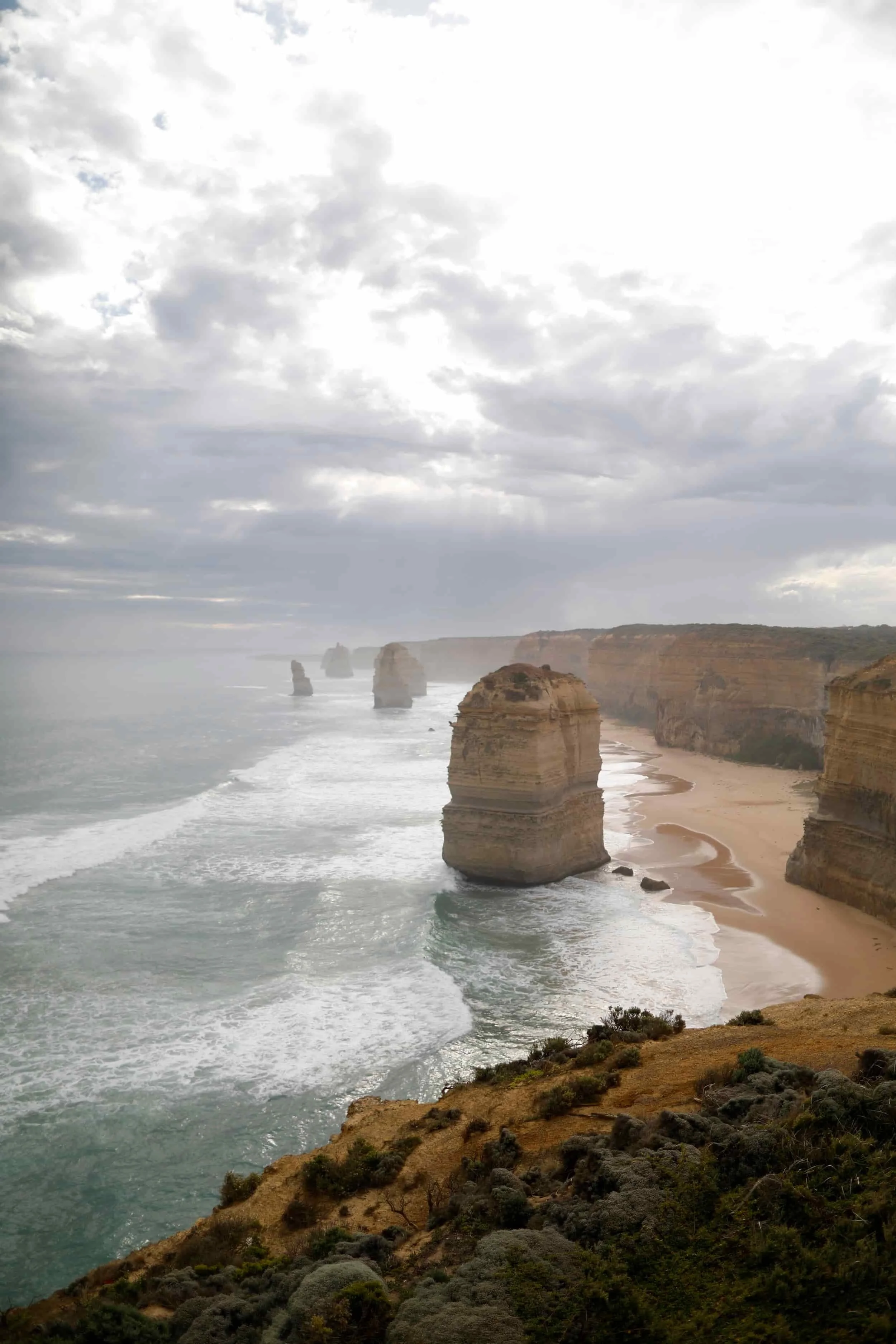 Overcast sky over the ocean with limestone stacks and rocky cliffs along a sandy beach, with coastal vegetation in the foreground.