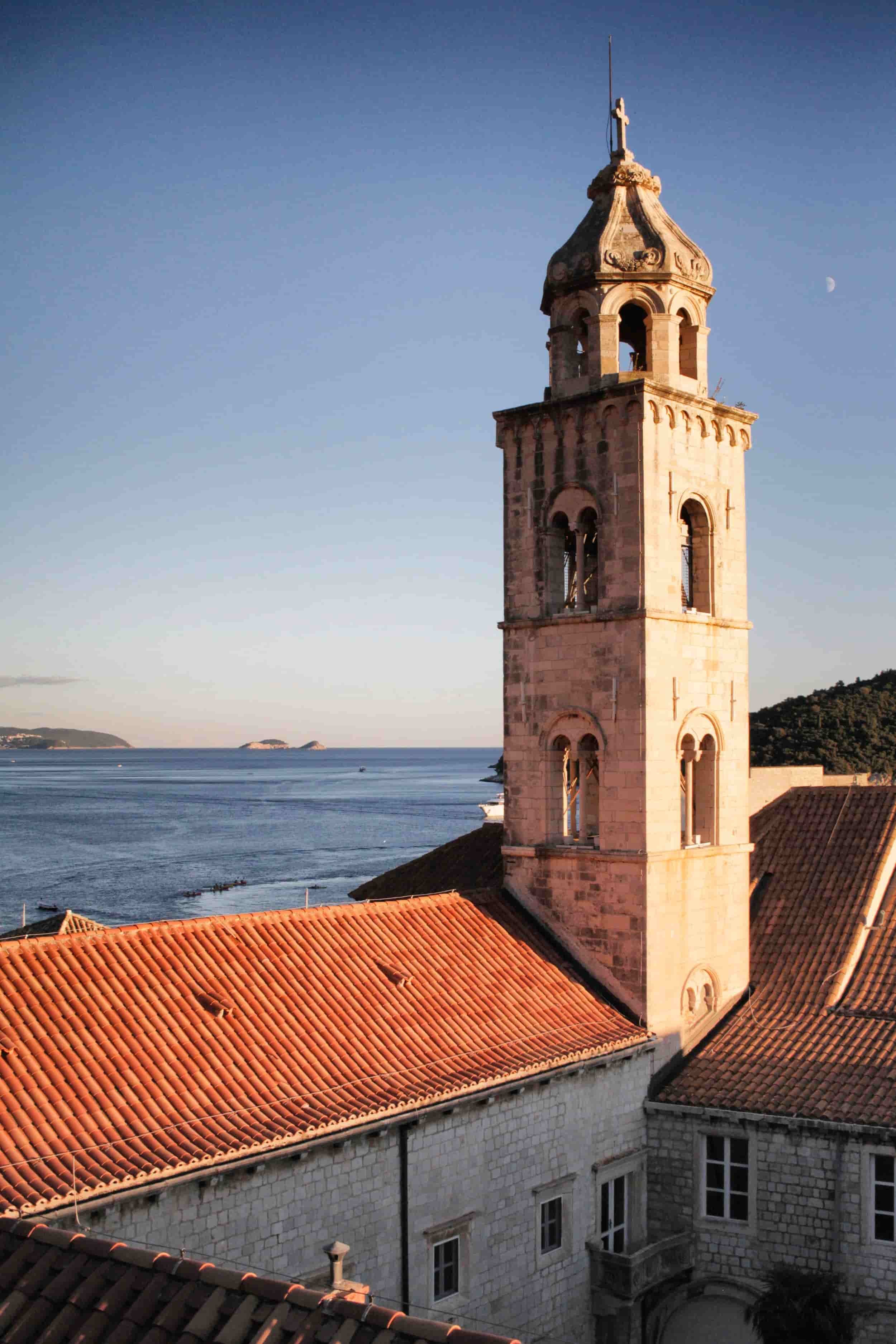 A historic stone church tower with a bell window under a shocked dome and cross on top, seen at sunset, against a blue sky with a crescent moon and ocean in the background.