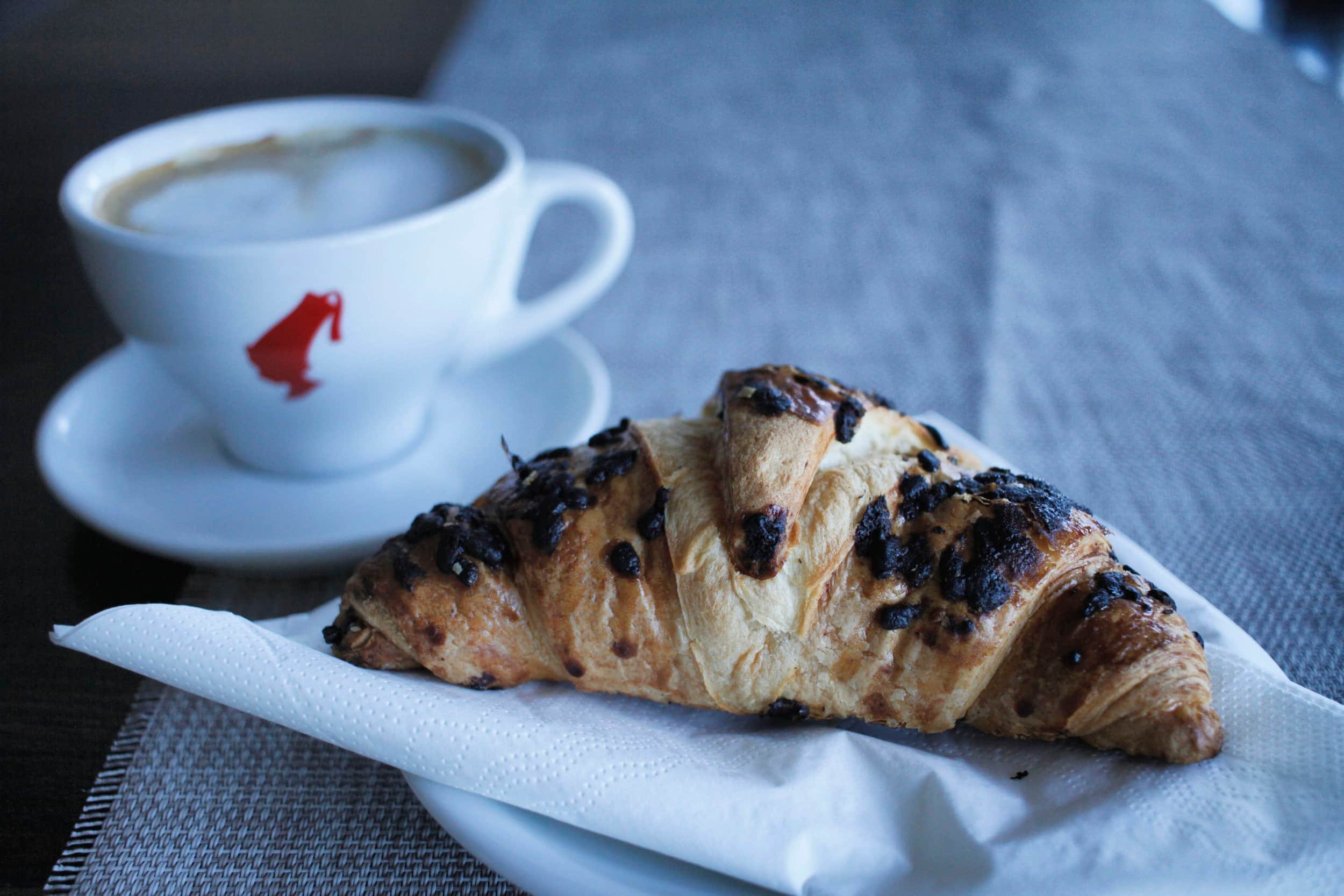 A white cup of coffee or cappuccino with frothy milk sits on a plate with a croissant topped with chocolate bits, on a table with a white paper napkin and a gray placemat.