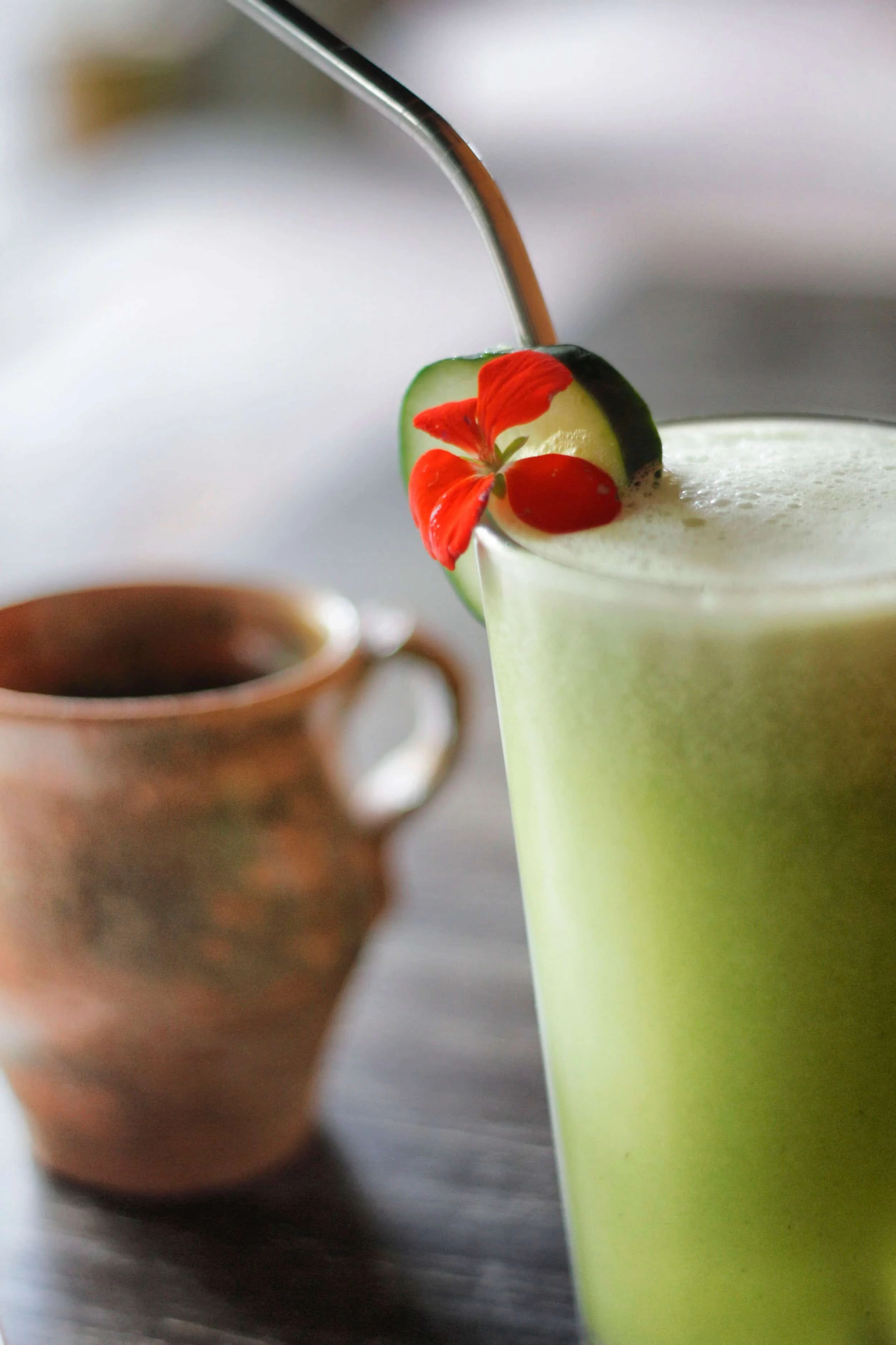 A close-up of a light green drink garnished with a cucumber slice and red edible flower, with a copper mug in the background.