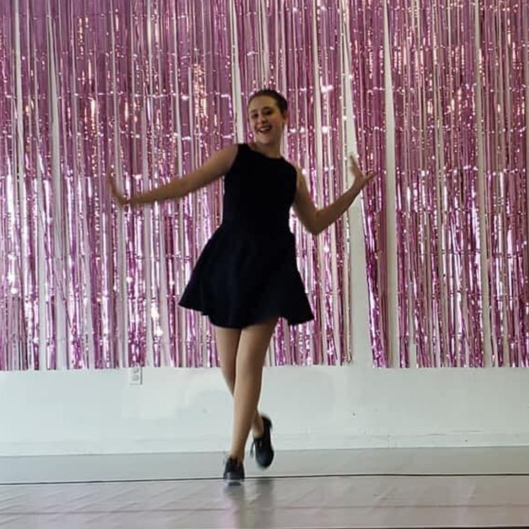 A young tap dancer wearing a black sleeveless dress and tap shoes performs in a dance studio in front of a pink shimmering backdrop.