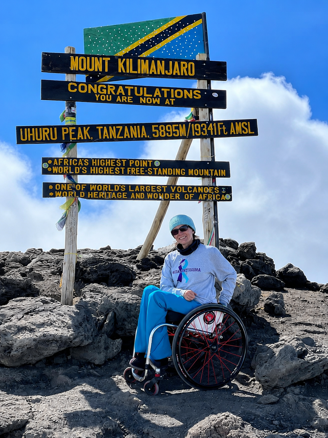 A woman in a wheelchair at the summit of Mount Kilimanjaro, Tanzania, smiling and wearing sunglasses and a light blue hat. Behind her is a sign indicating the location as Uhuru Peak, the highest point on Kilimanjaro, with details about the elevation and mountain's significance.