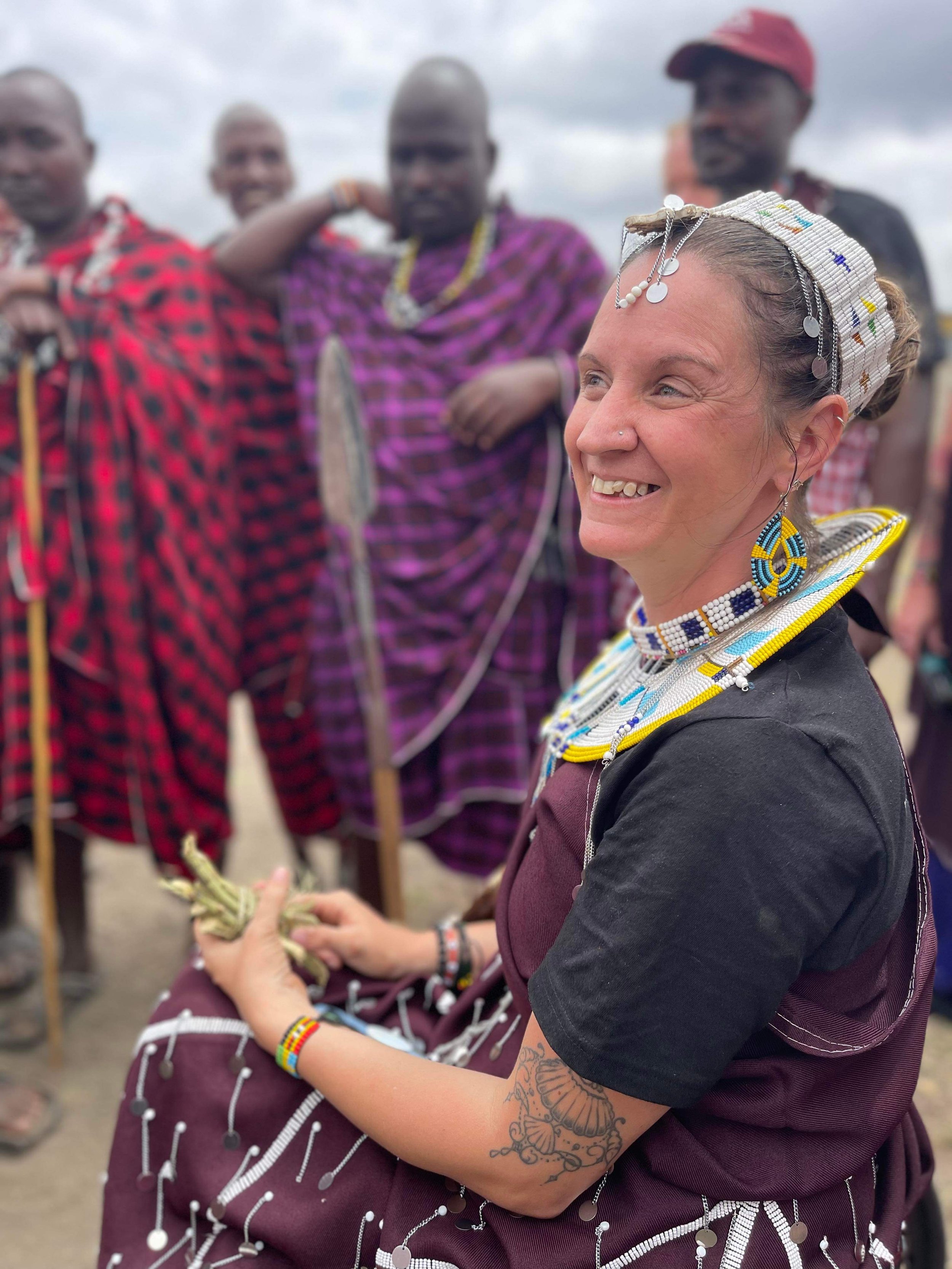 A woman in traditional Maasai attire, wearing beaded jewelry and a beaded headpiece, smiling as she participates in a cultural exchange with Maasai men dressed in colorful shuka garments, outdoors with a cloudy sky.