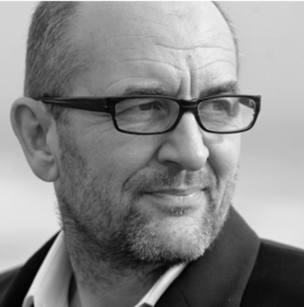 Black and white close-up of a middle-aged man wearing glasses, with a slight smile, short hair, and a beard, dressed in a suit jacket.
