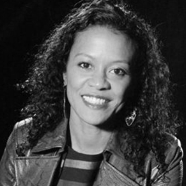 Black and white portrait of a smiling woman with curly hair wearing a leather jacket and striped shirt.