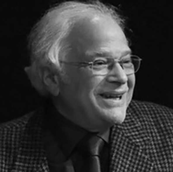 Black and white photo of a smiling elderly man with white hair, glasses, wearing a checkered blazer and dark shirt with a tie.