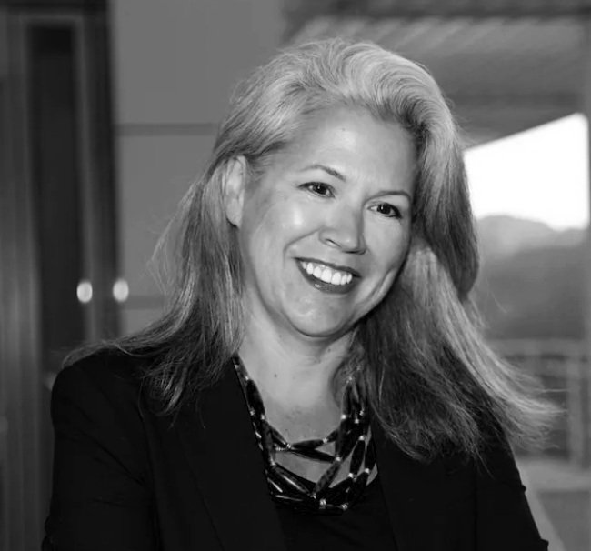 A woman with long, wavy blonde hair, smiling, wearing a black blazer and a patterned necklace, indoors with a window in the background.