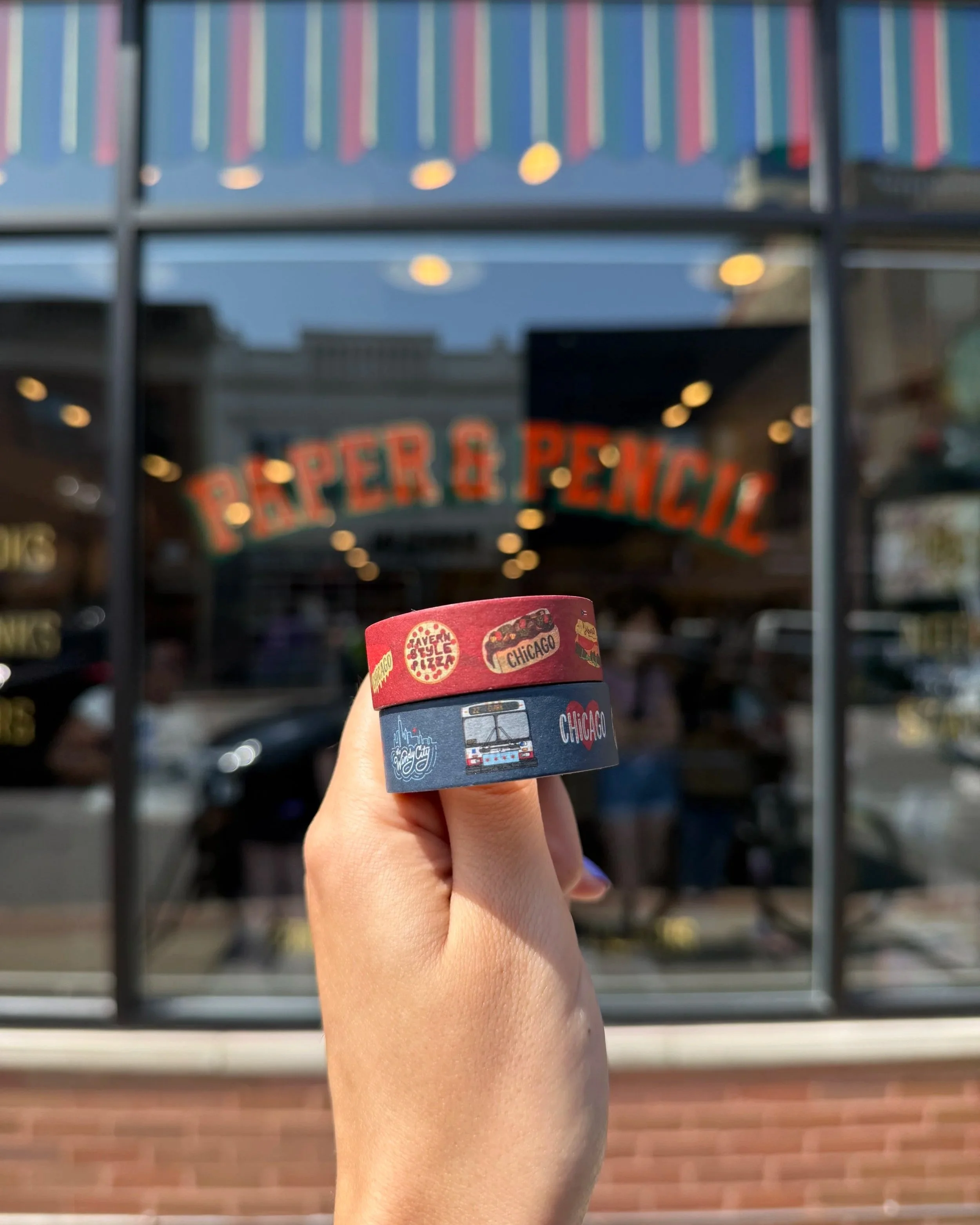 Chelsie holds up two rolls of Chicago style washi tape in front of the Paper & Pencil storefront. One roll is red with Chicago food illustrations and the other roll is navy with other Chicago illustrations.