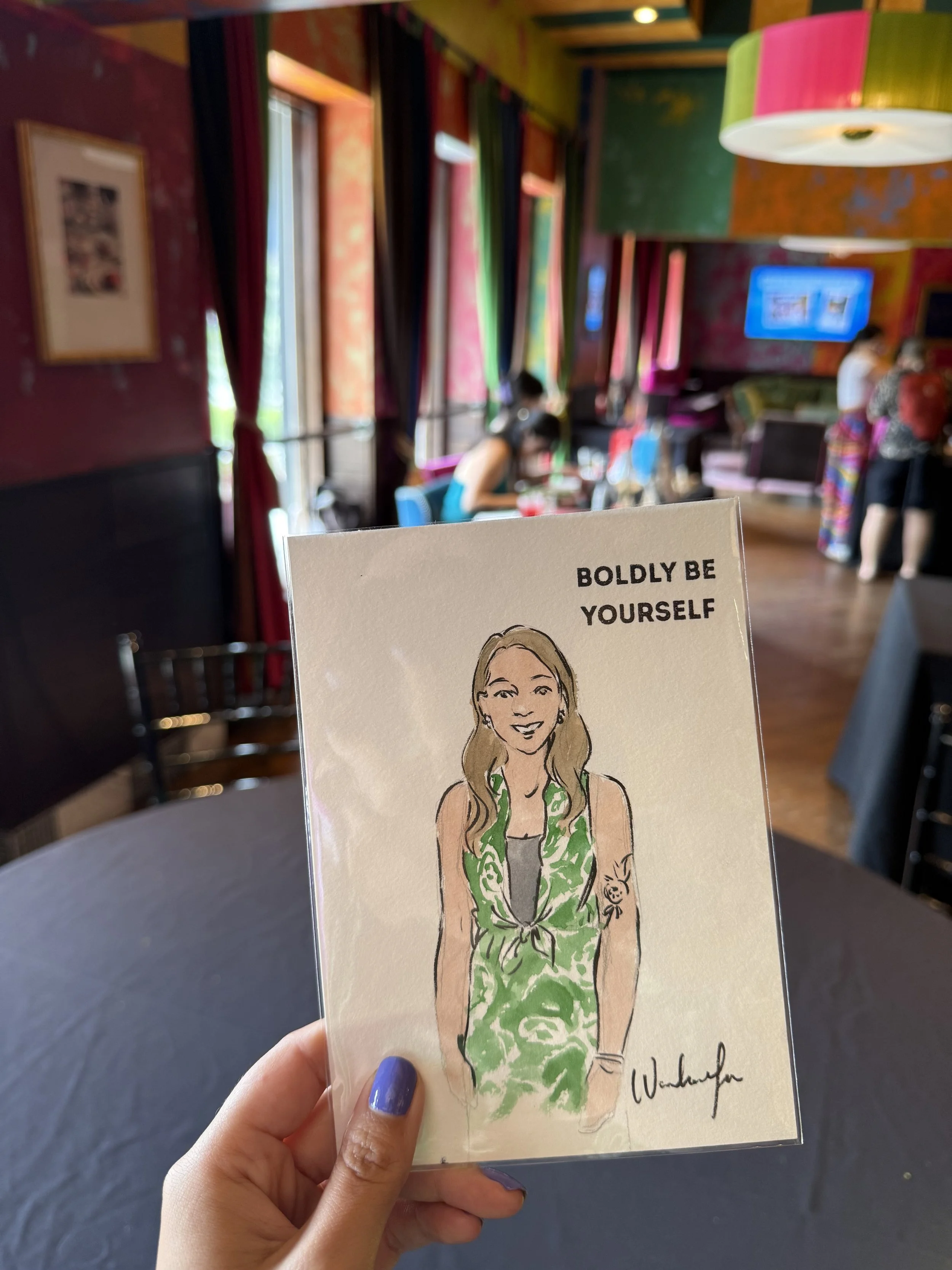 Chelsie is holding a small live-painted portrait that showcases her vibrant green and cream pineapple outfit and highlights her pomegranate tattoo. In the background there is a table where the artist sits painting additional live portraits.