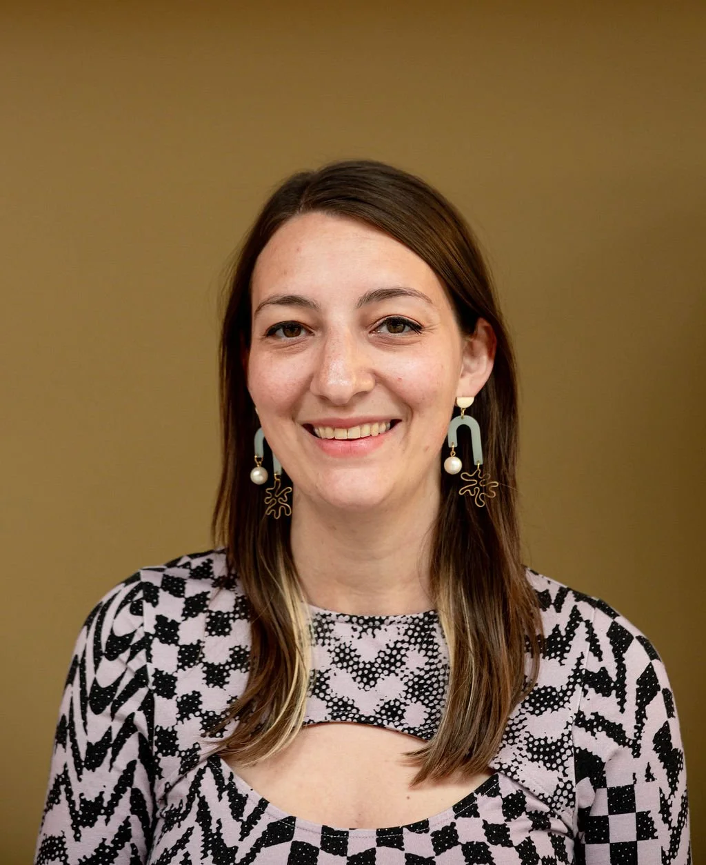 Headshot of Lettering Artist and Chicago Souvenir Designer Chelsie Tamms. Chelsie is smiling in a pink and black checkered dress with fun earrings and straight medium brown hair.