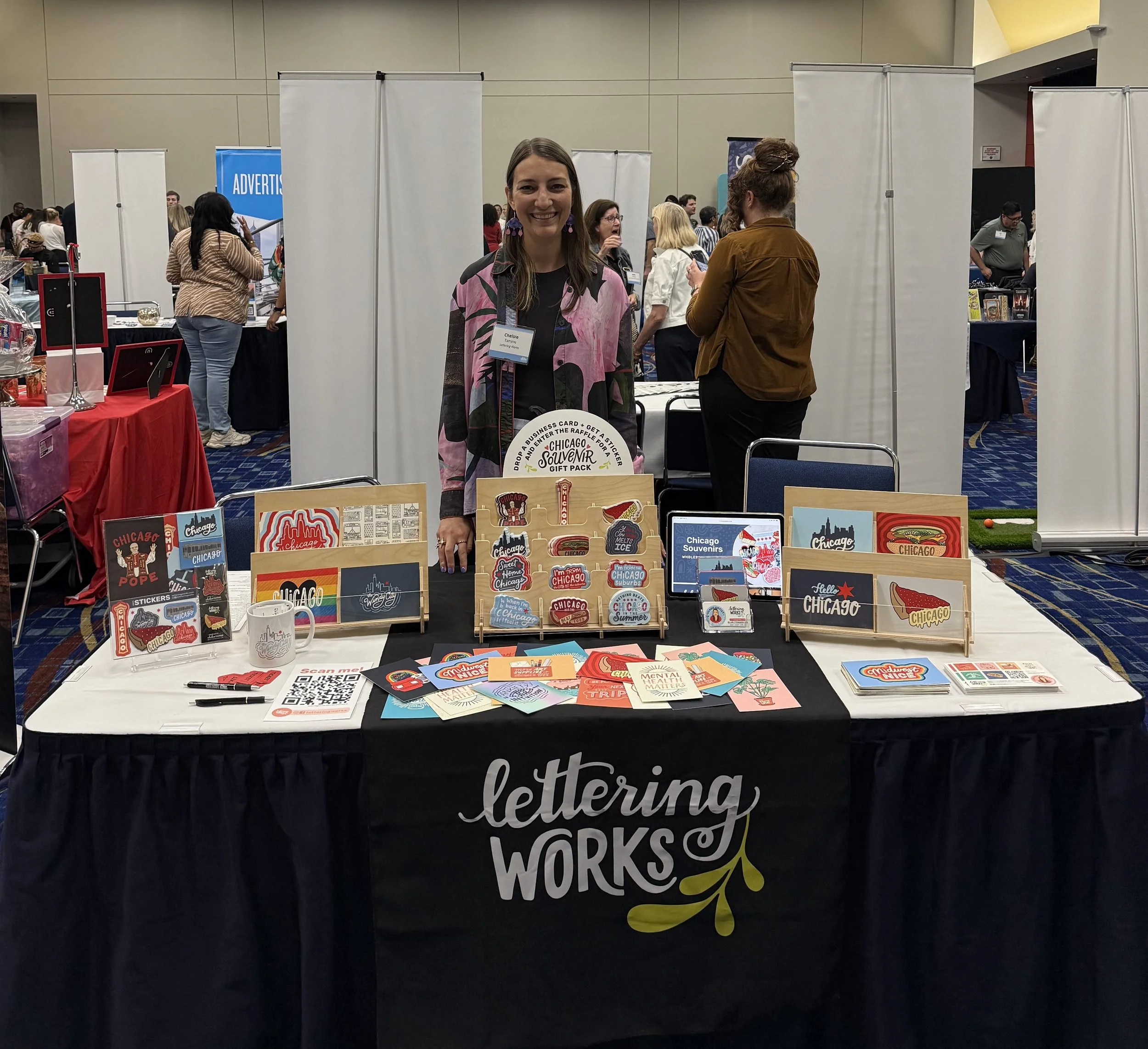 Artist Chelsie stands behind her trade show booth which features stands of vibrant Chicago stickers and Chicago postcards as well as promotional cards. The table is covered in a black table cloth with a table runner that displays Lettering Works logo