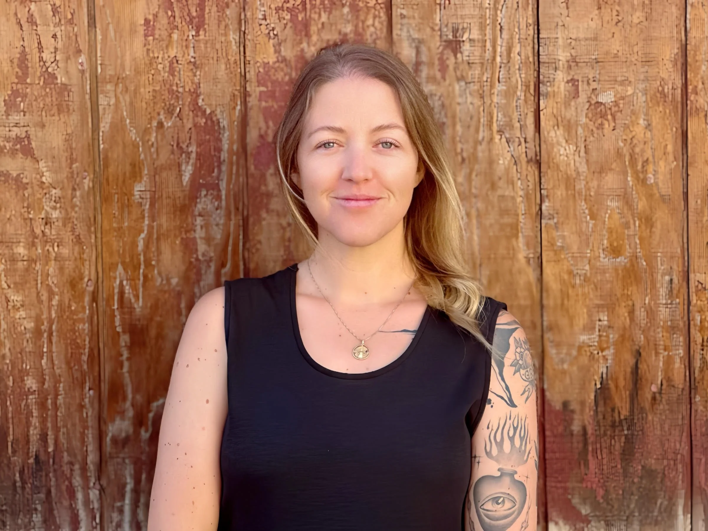 A young woman with long blonde hair posing outdoors against a wooden fence. She is wearing a black sleeveless top and has tattoos on her left arm and a small tattoo on her chest. She is smiling slightly and looking at the camera.