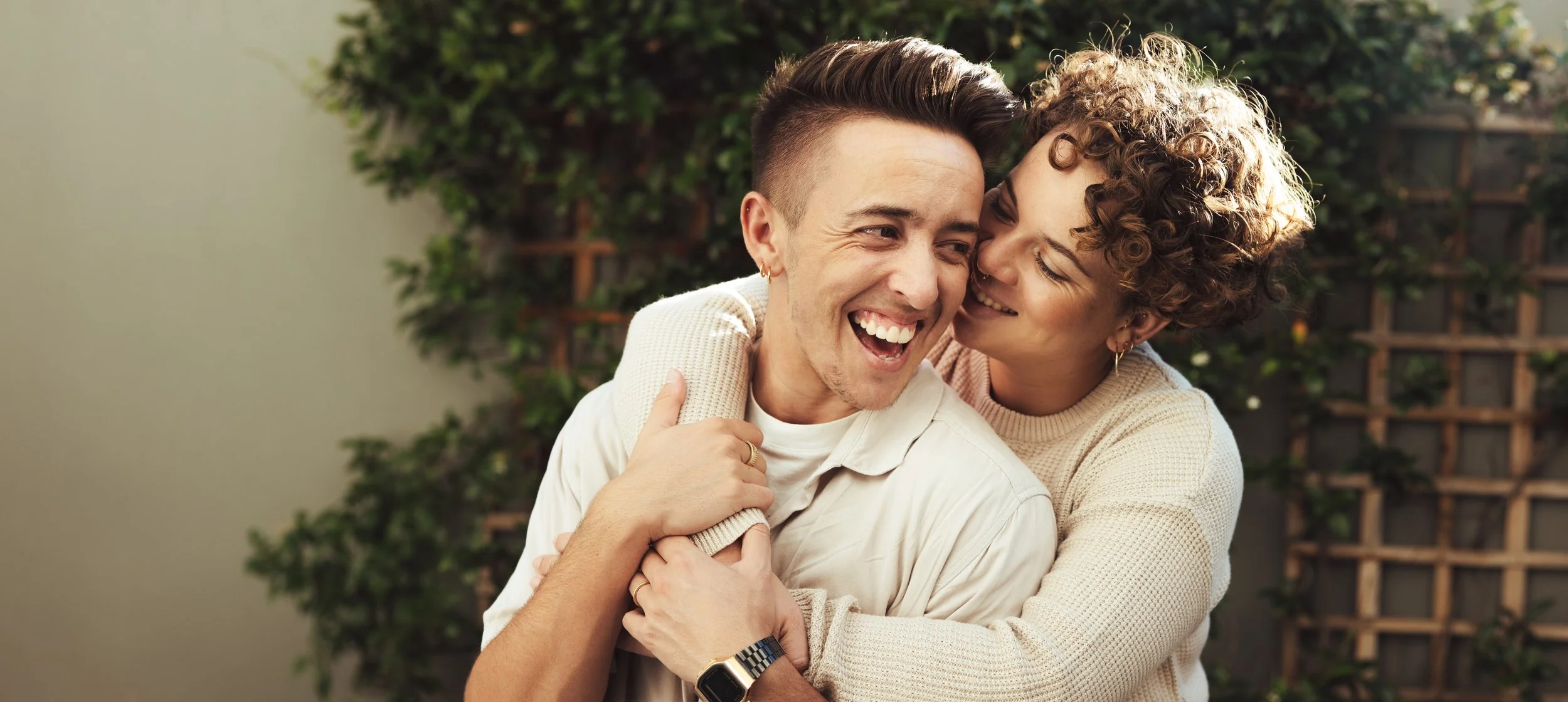 A happy couple embracing and laughing outdoors with greenery in the background.