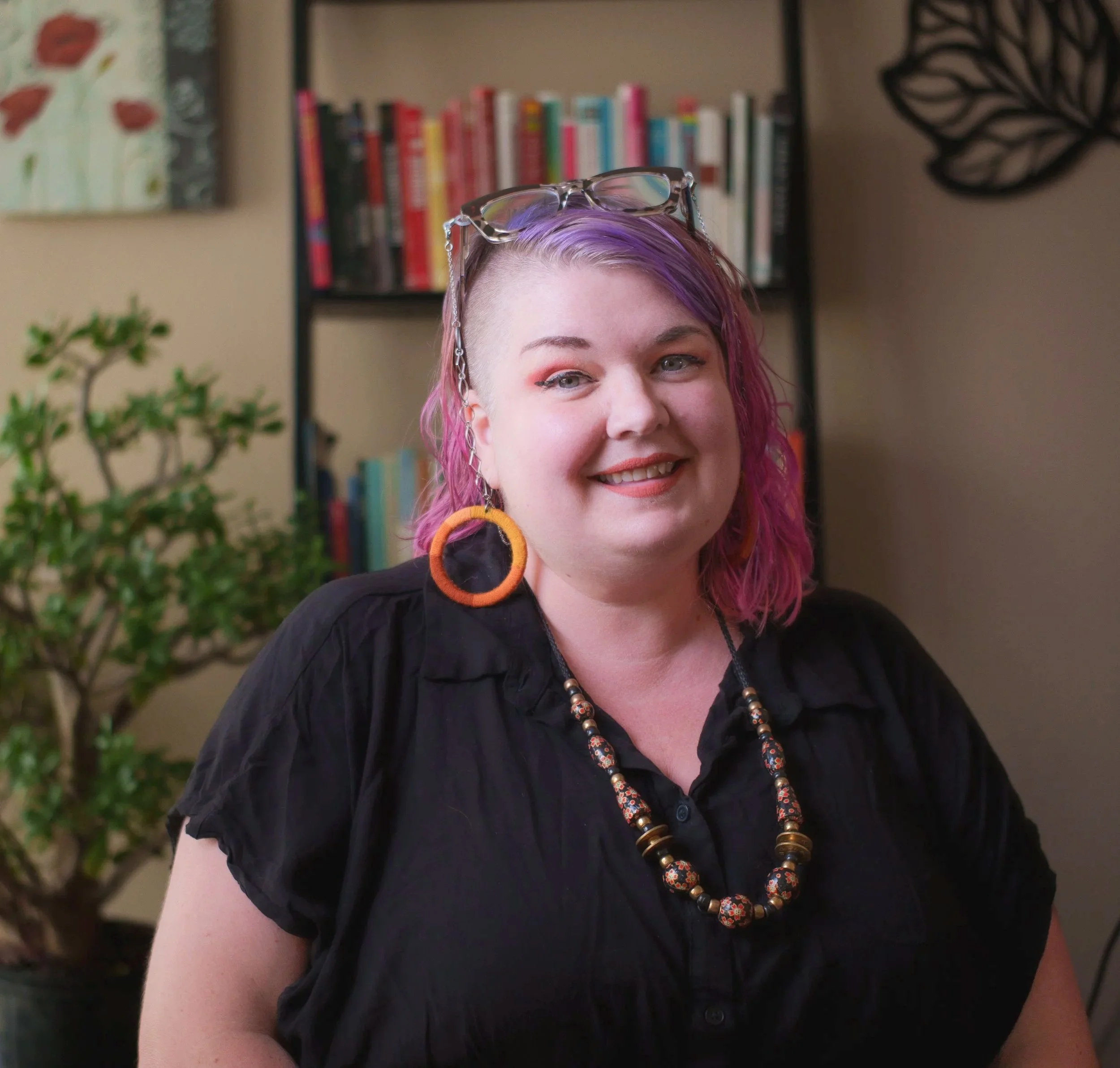 A person with pink hair, partially shaved on one side, smiling and wearing glasses on their head, a black top, and large orange earrings with a beaded necklace. In the background, there are books on a shelf,  paintings and plant.