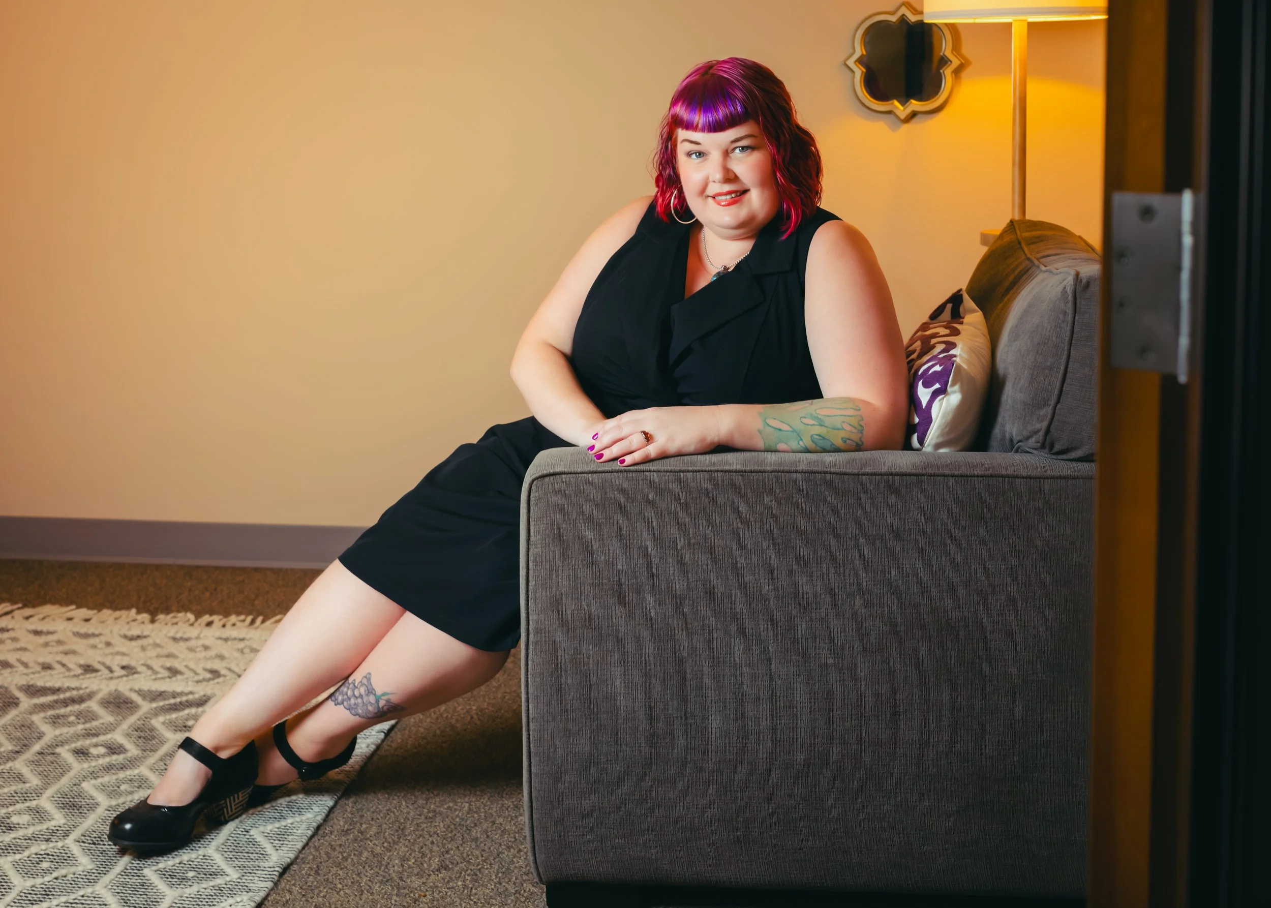 A non binary person with purple and pink hair sitting on the arm of a gray sofa, smiling, with a tattoo on their left leg and wearing a black sleeveless top, in a warmly lit room with a yellow wall, a decorative mirror, and a patterned rug.