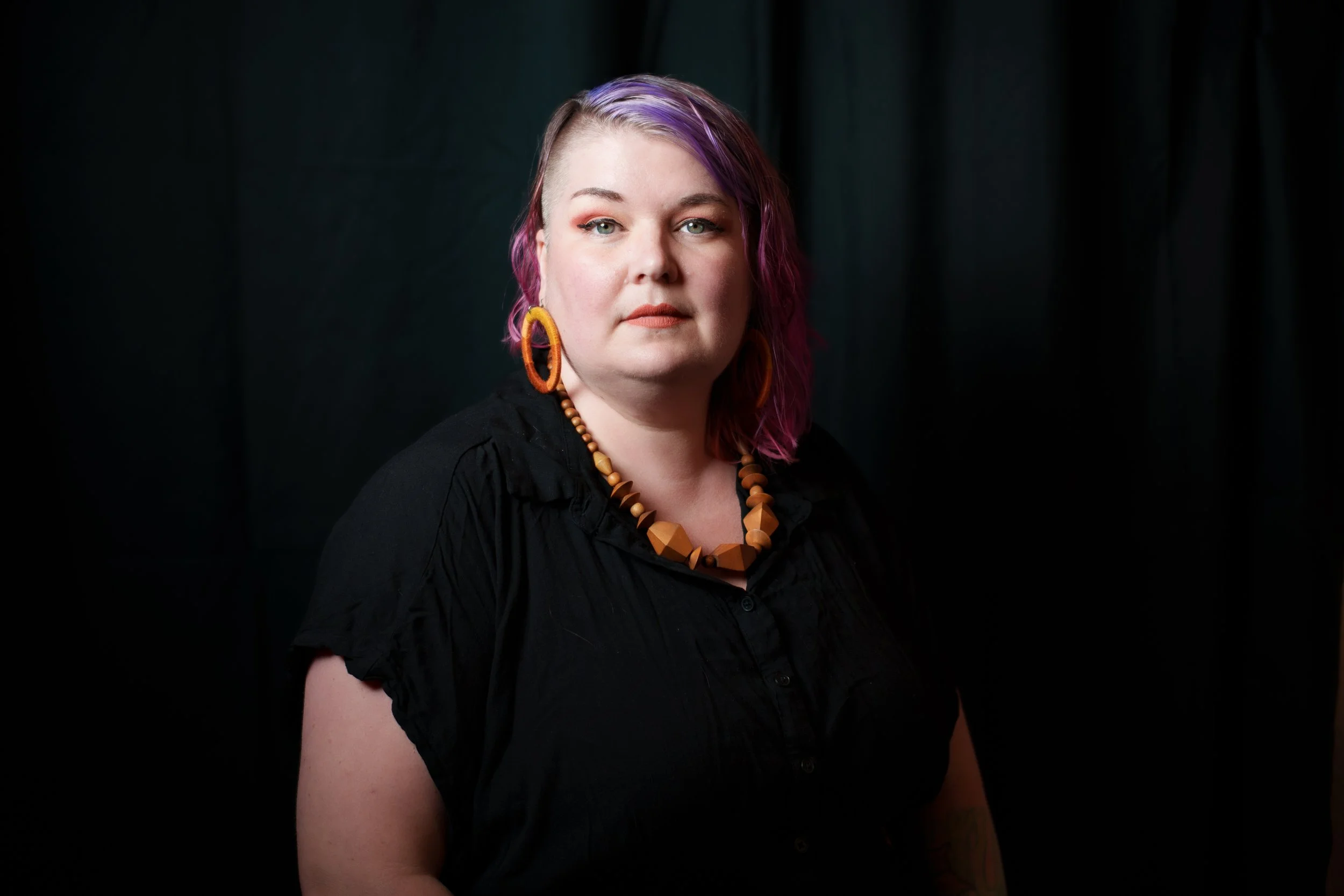 A person with short purple and pink hair, wearing orange earrings and a beaded necklace, dressed in a black top, against a dark background.