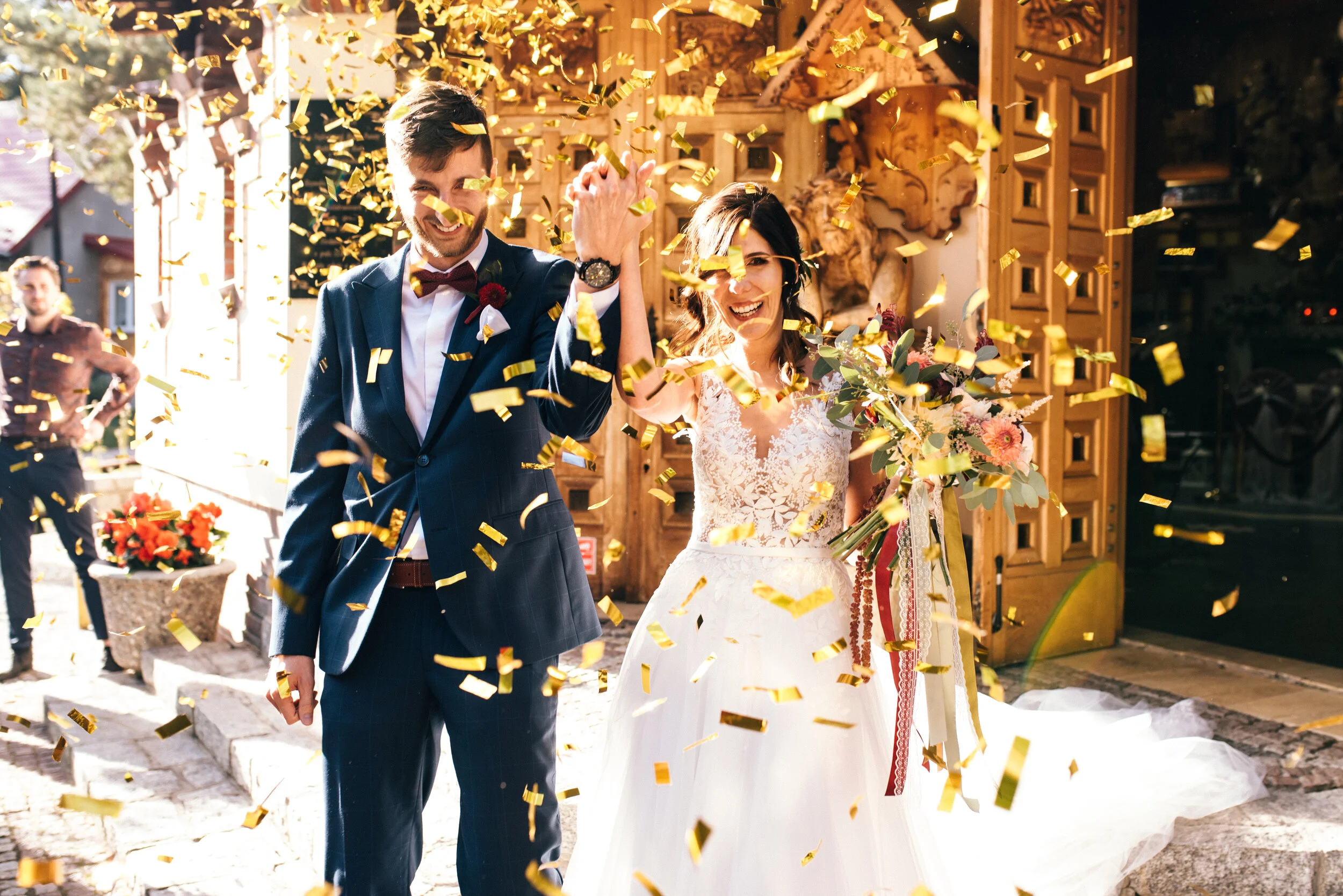A newlywed couple smiling and celebrating as they walk through falling gold confetti, holding hands, with a wooden door and outdoor setting in the background.