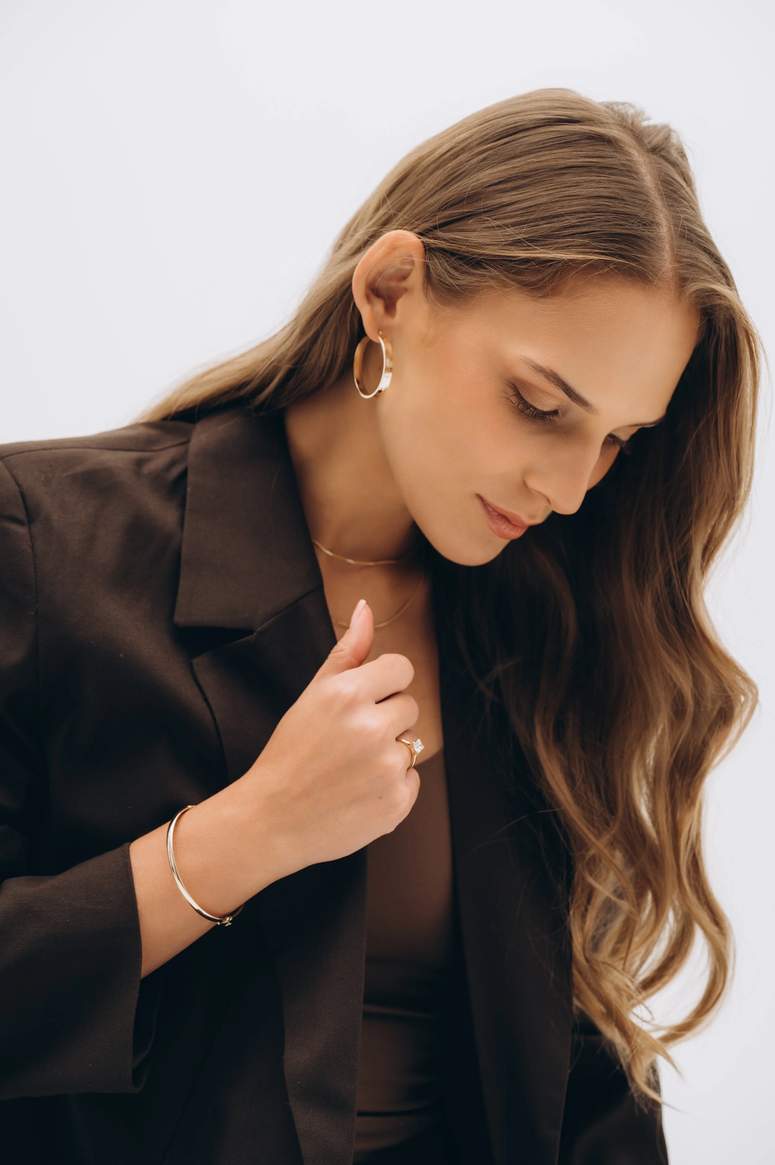A woman with long, wavy brown hair, wearing gold hoop earrings, a gold necklace, a ring, and a bracelet, dressed in a black blazer and top, looking down.