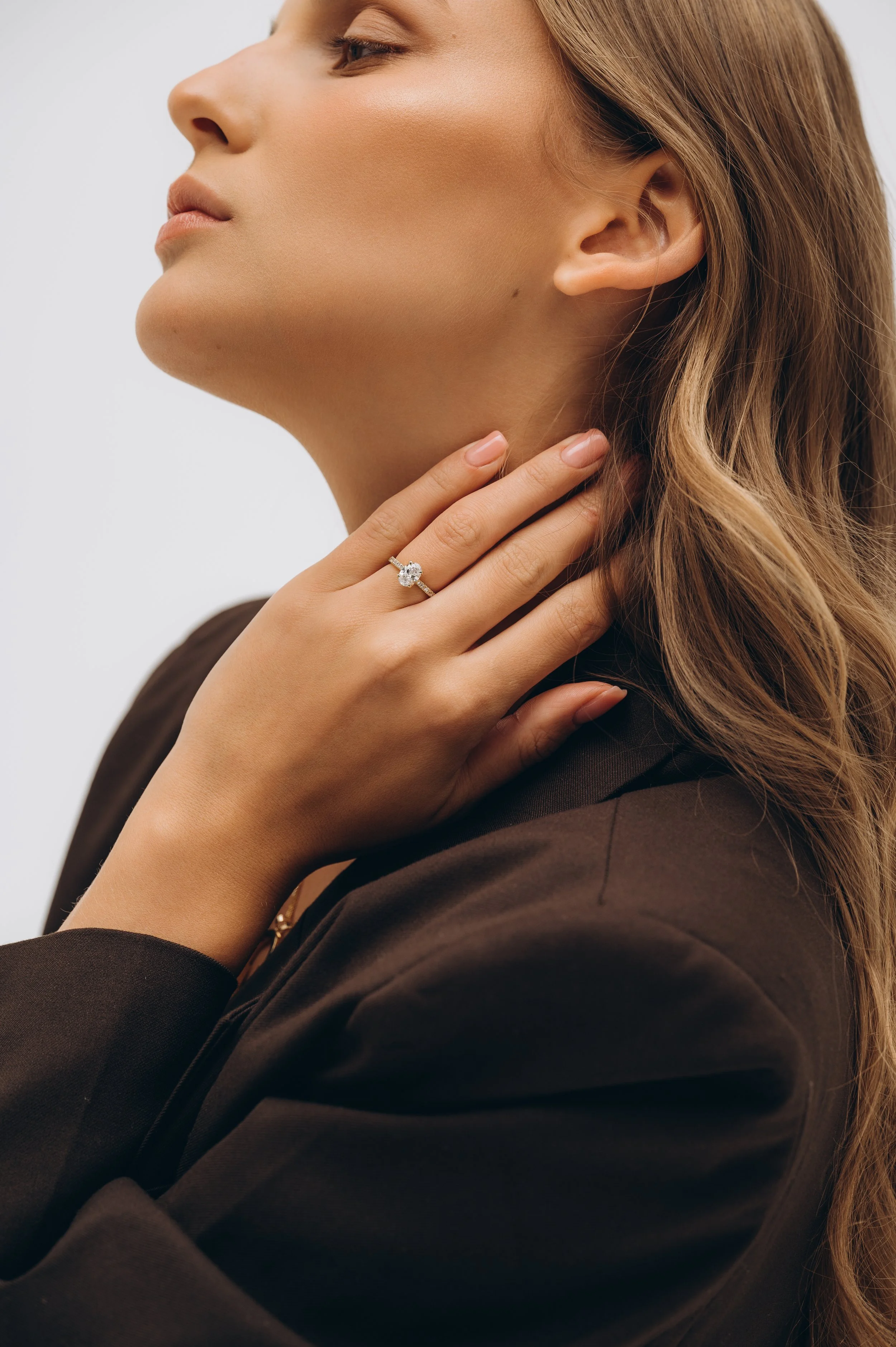 A woman with wavy blonde hair showing her profile, wearing a dark blazer, and a diamond engagement ring on her finger, touching her neck.