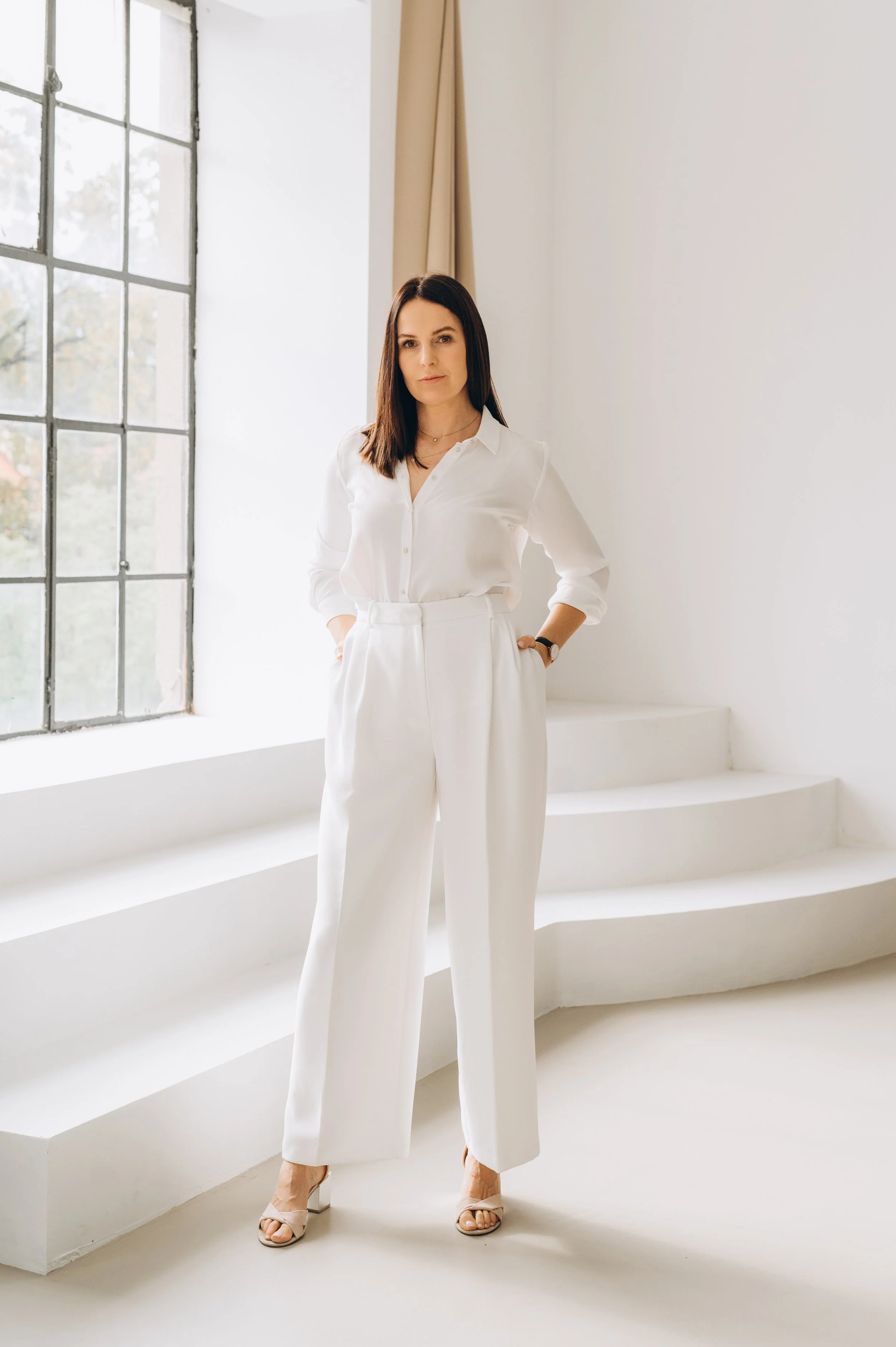 A woman with brown hair, wearing a white button-up shirt, white wide-leg pants, and beige heels, stands inside a minimalistic white room with large windows.