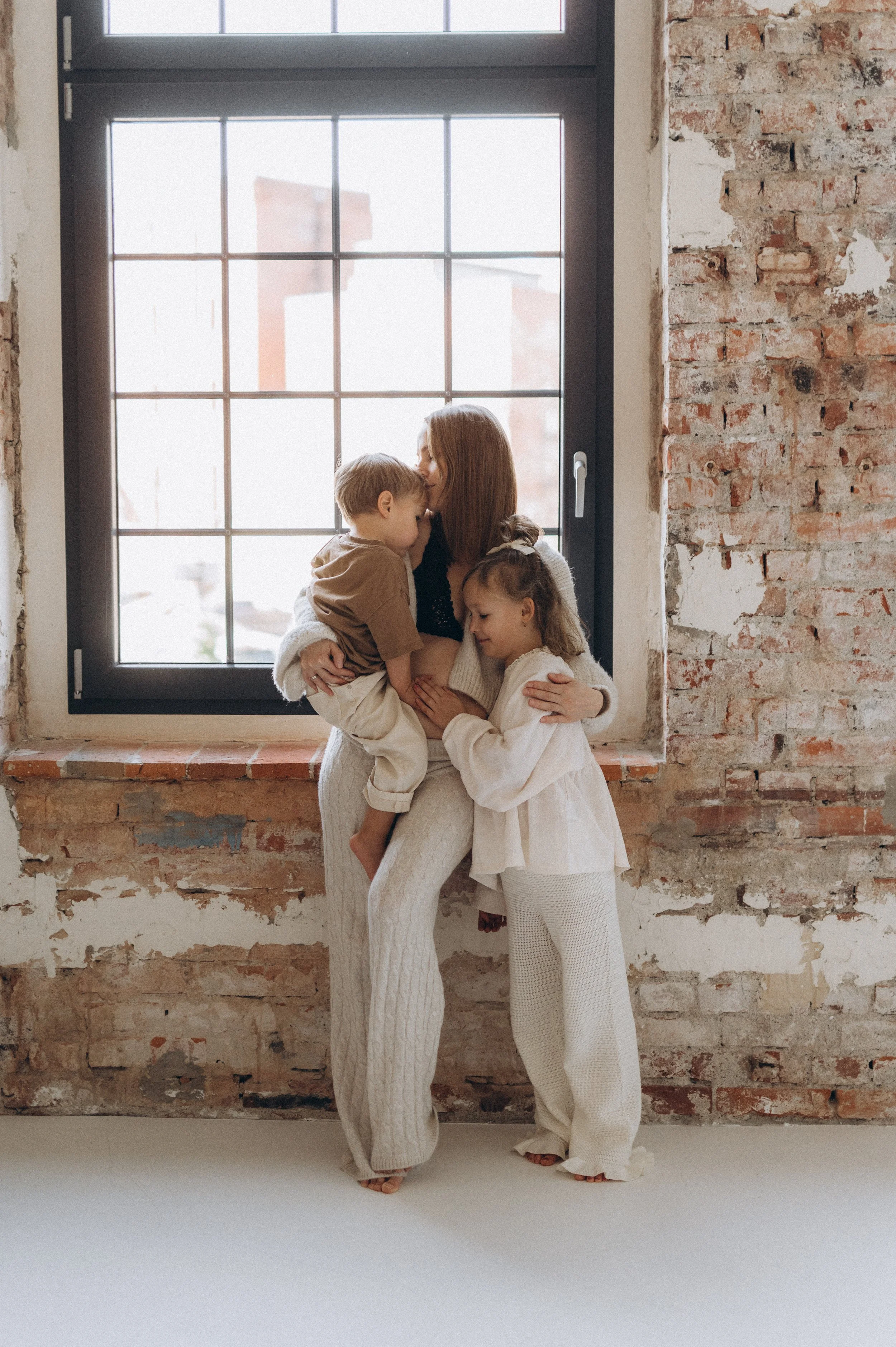A woman hugging two children by a large window with brick walls.