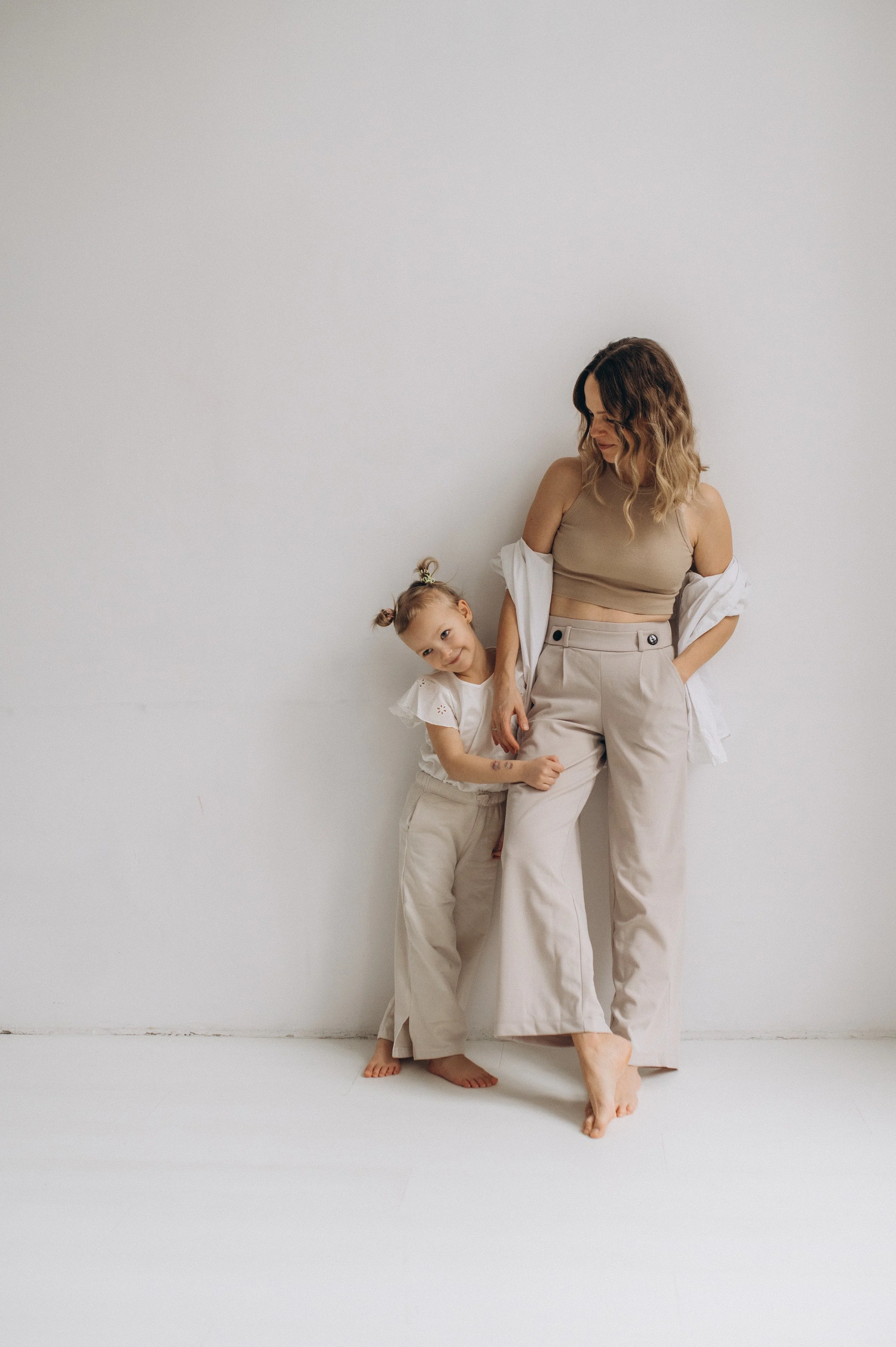 A woman with shoulder-length wavy hair in beige pants and a beige crop top stands barefoot against a white wall, with a young girl in matching beige pants and a white blouse leaning on her and smiling.