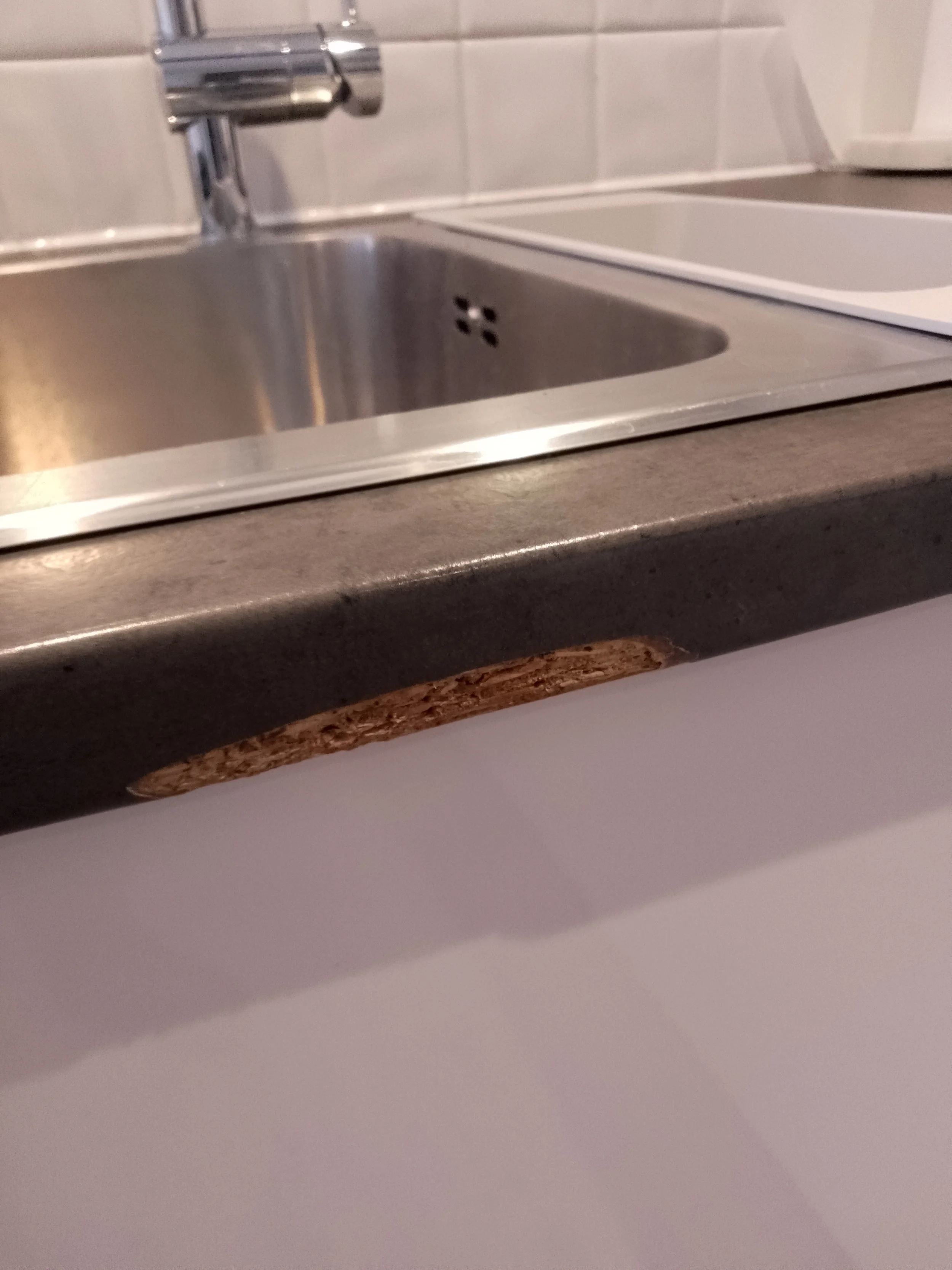 Close-up of a kitchen worktop with a damaged, chipped edge revealing the particleboard underneath, stainless steel sink, and white tiled wall.