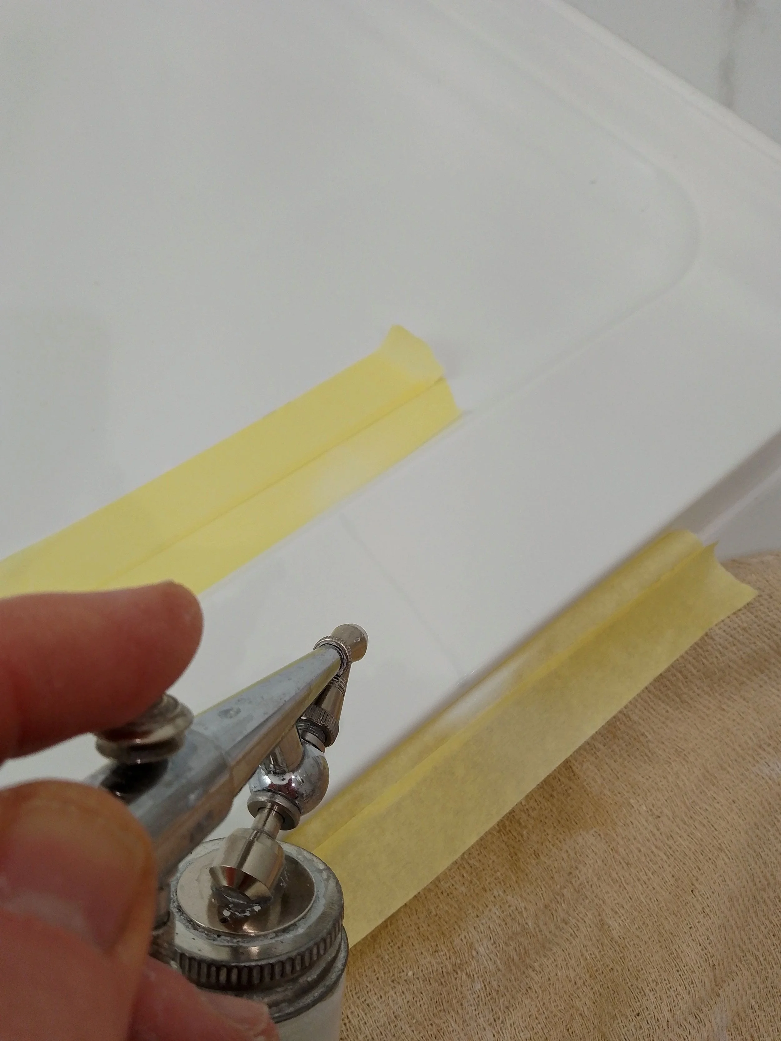 A person using an airbrush spray tool to paint or finish a white surface, with masking tape applied along the edges.