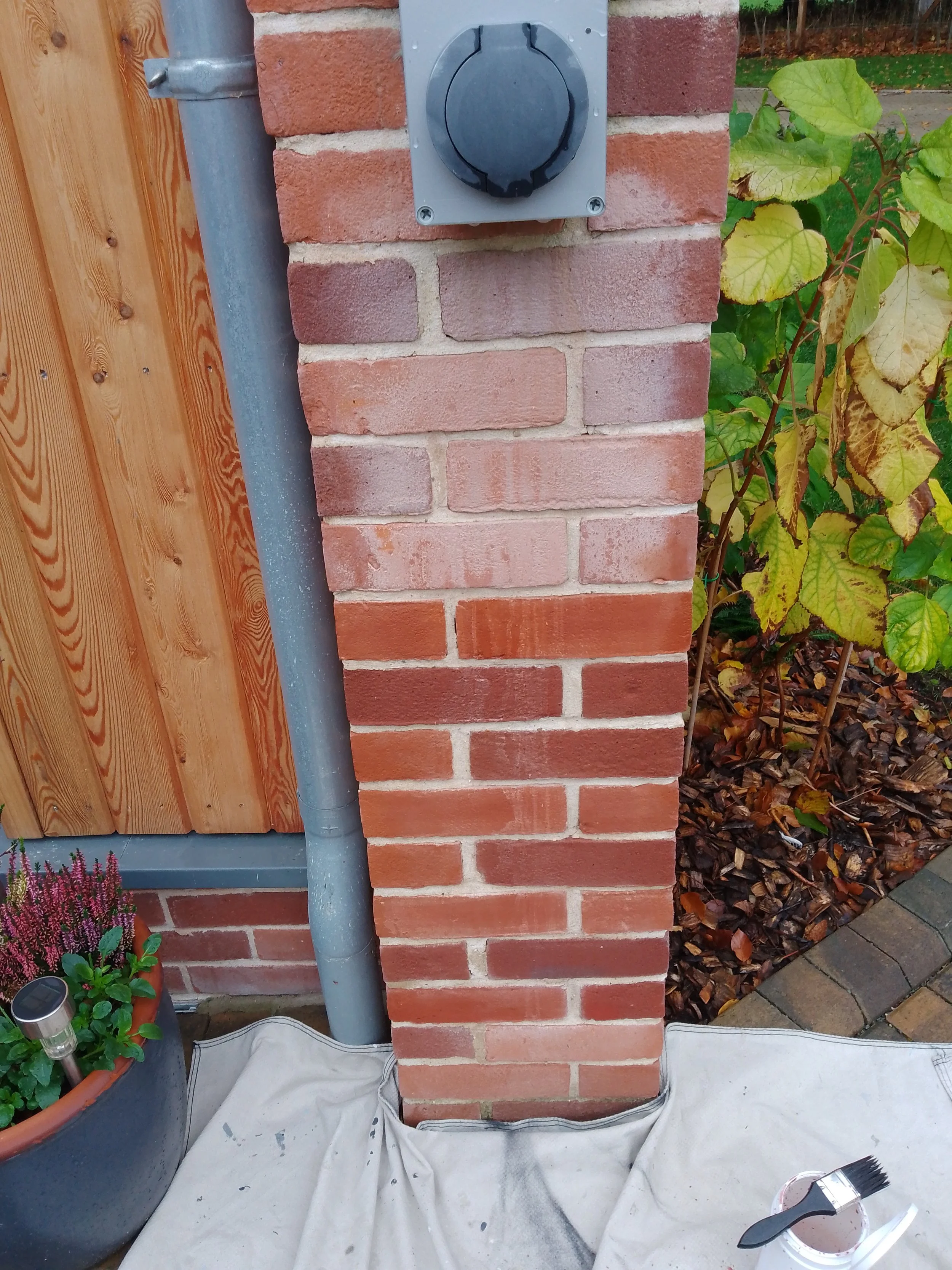 Close-up of a brick pillar with an electrical box and pipe, surrounded by garden plants and a wooden fence, on a paved patio.
