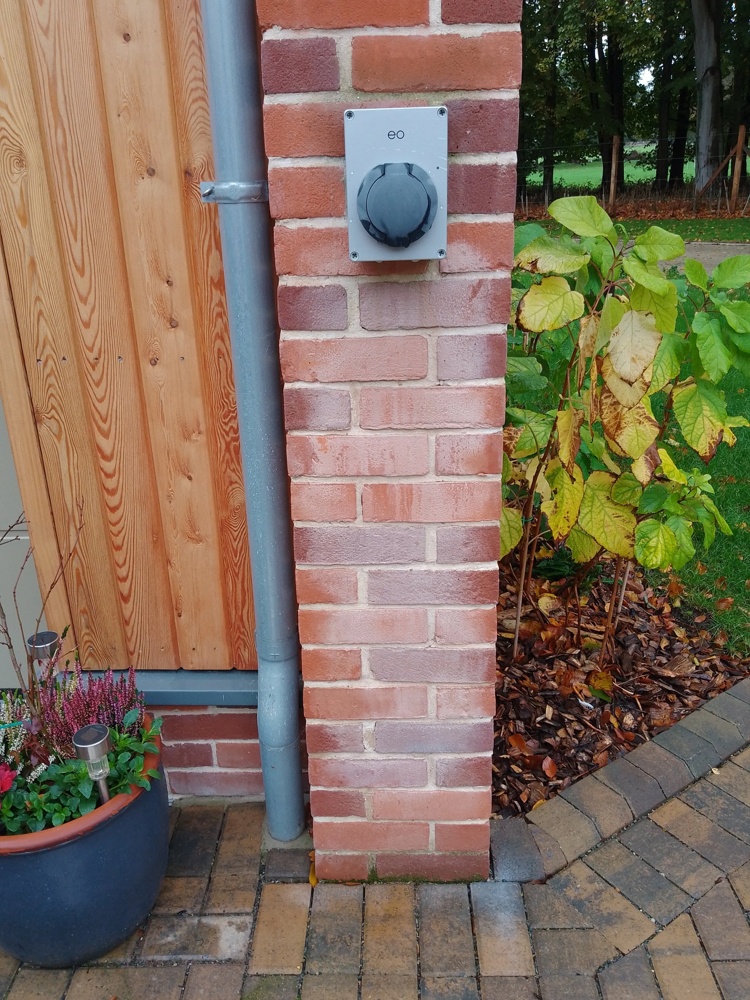 Close-up of a brick pillar with a black outdoor electrical outlet on a white mounting plate, flanked by a metal downspout on the left and a wooden wall on the left, with a potted flower on the ground and some green foliage and trees in the background.