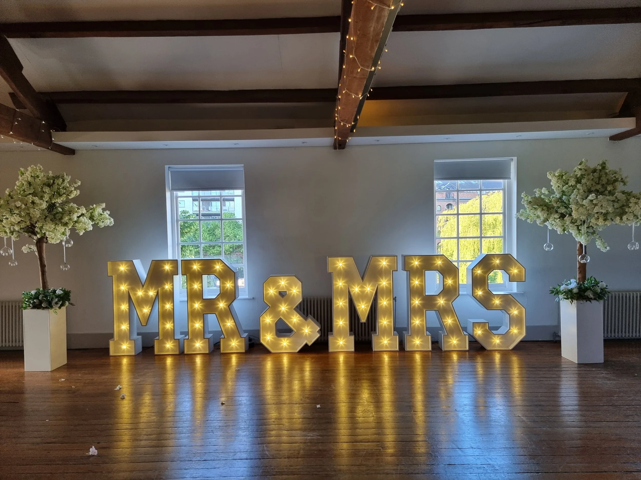 Decorative lit signs reading 'MR & MRS' with two white flower trees on either side in a room with wooden floors and windows showing greenery outside.