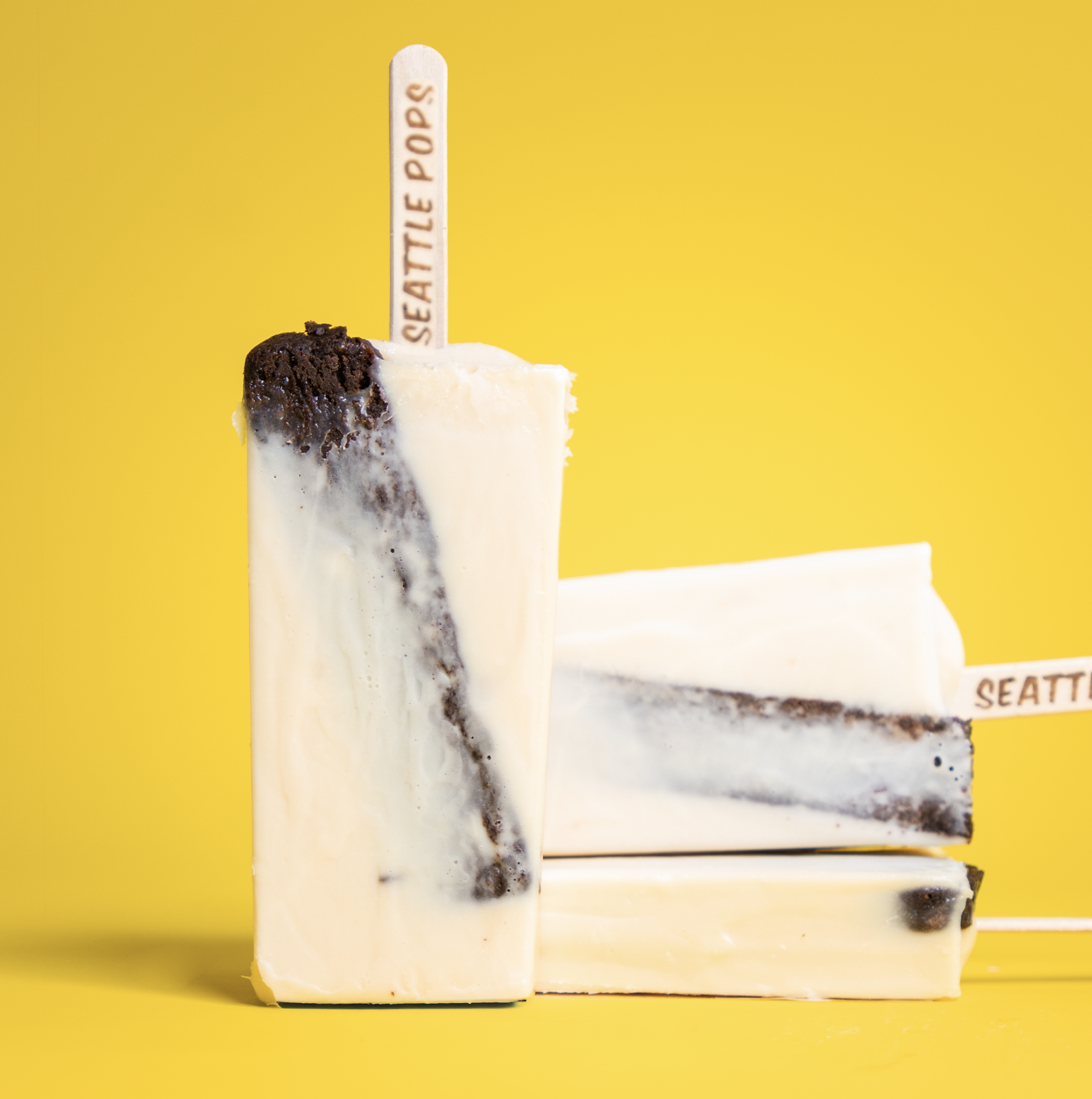 Two ice cream bars with a vanilla coating and chocolate swirls on a yellow background, one standing upright and the other lying flat, each with a wooden stick labeled 'SEATTLE POPS'.