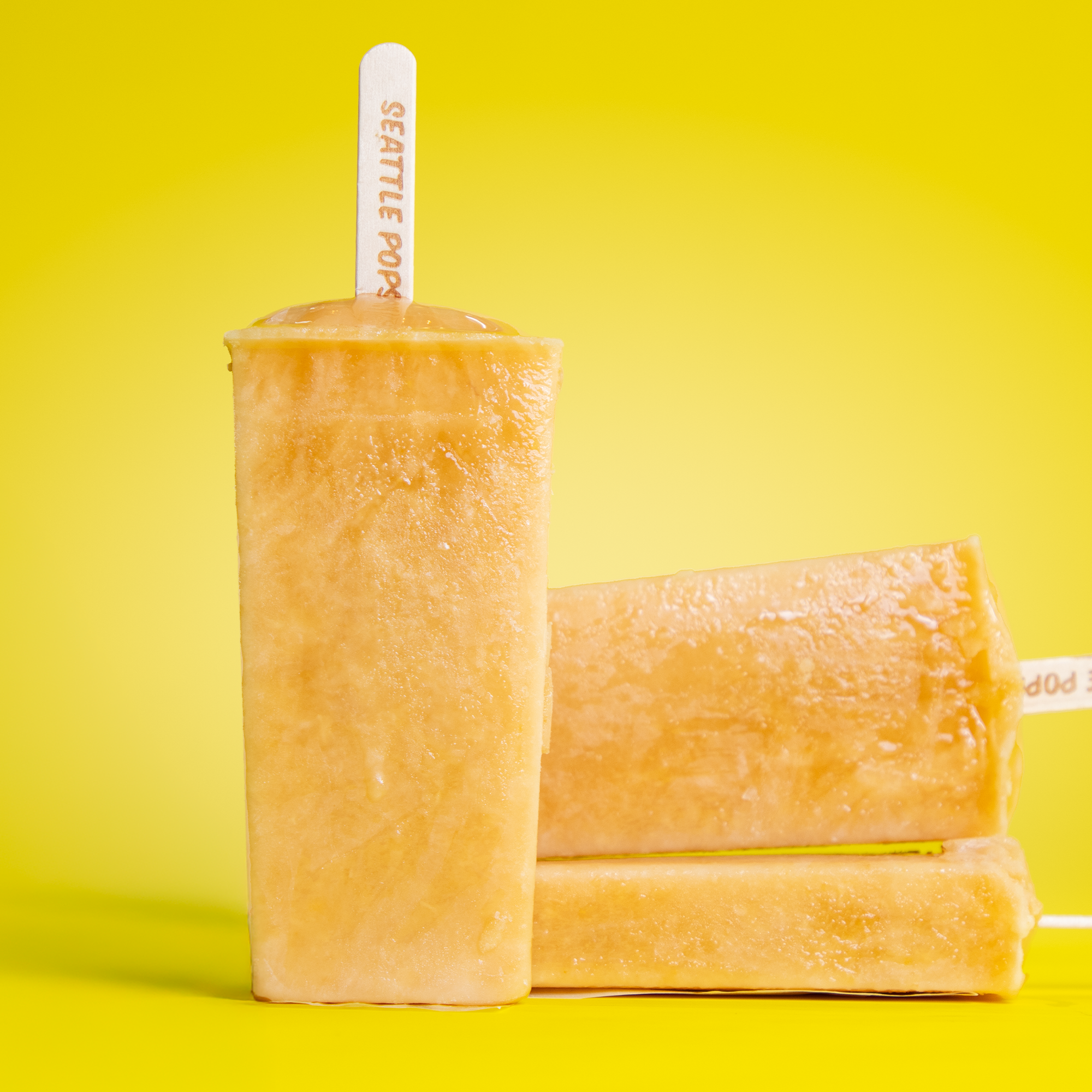 Two rectangular frozen popsicles with a yellow-orange hue, one standing upright and the other leaning on it, against a solid bright yellow background.