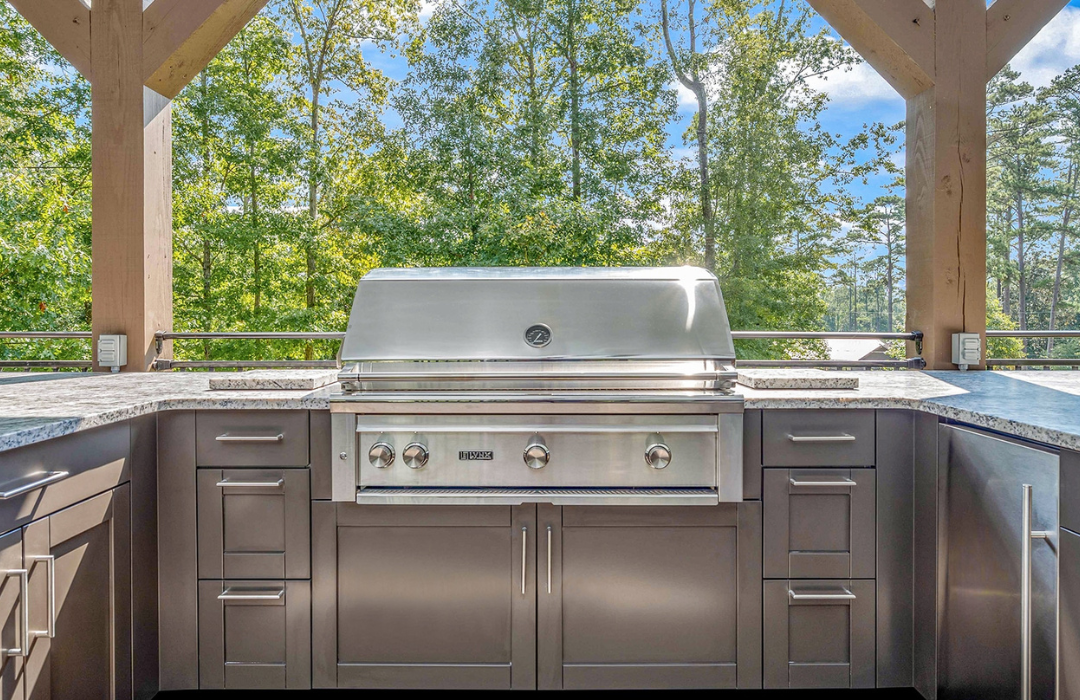 Outdoor kitchen under a timber pergola with stainless steel built-in grill, gray cabinets, and granite-look countertops surrounded by wooded views