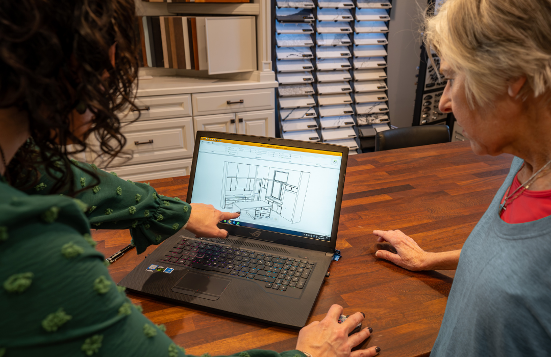 Two women reviewing a 3D closet design on a laptop at the HomeSource Design Center, with tile and cabinet samples in the background