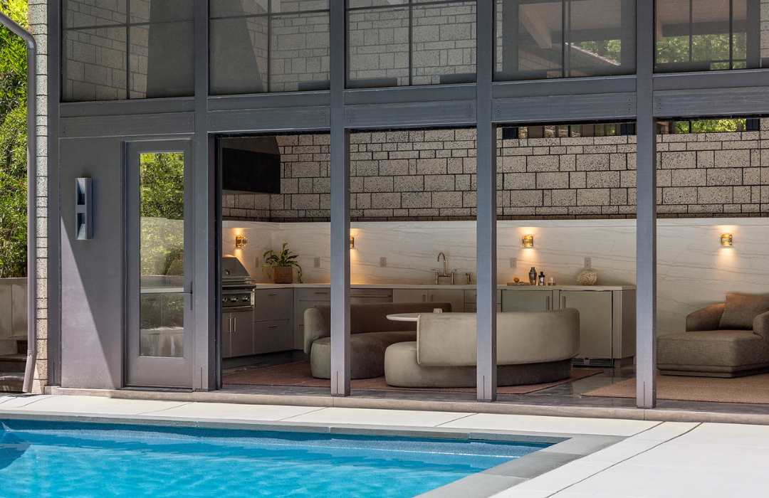 Enclosed outdoor kitchen and lounge area with gray cabinetry, marble-look backsplash, built-in grill, and poolside glass wall panels