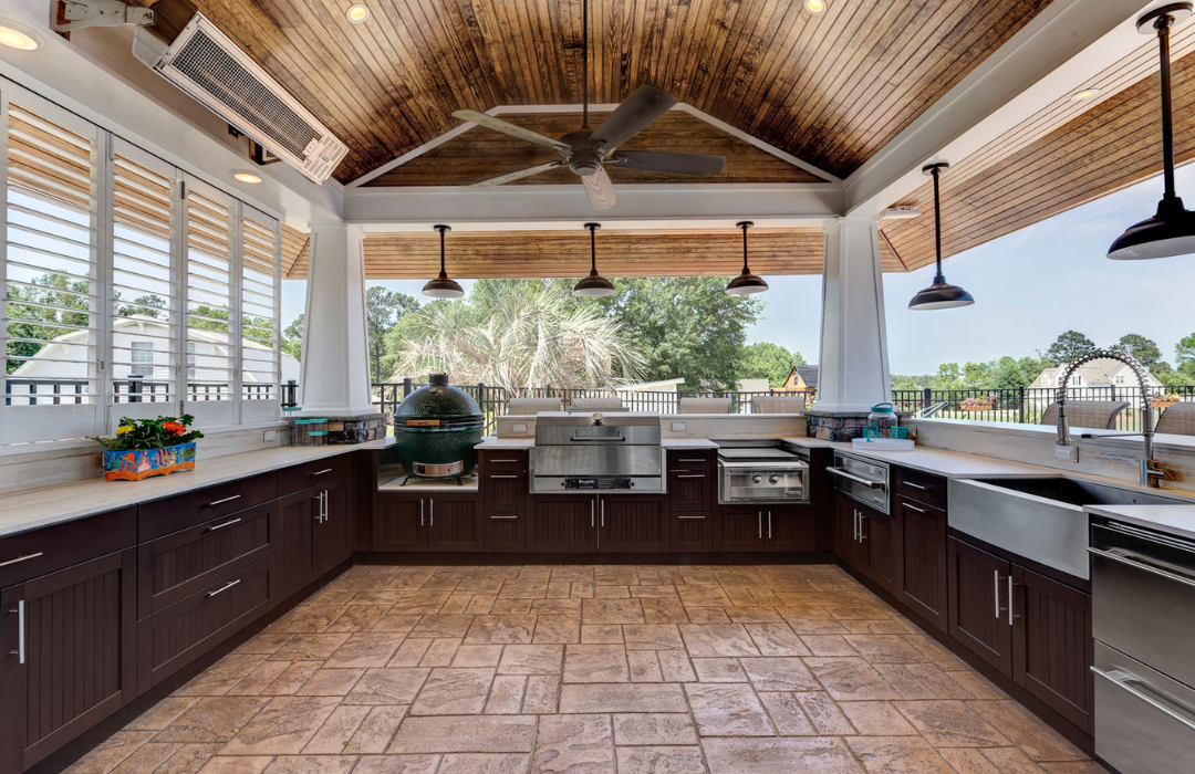 Large covered outdoor kitchen with dark wood cabinets, Big Green Egg smoker, built-in grill, farmhouse sink, and tile flooring