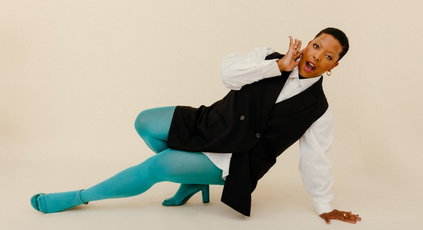 A woman with short hair wearing teal tights, teal high-heeled shoes, a white shirt, and a black vest, poses on the floor with one hand on the ground and the other hand near her ear, against a plain beige background.