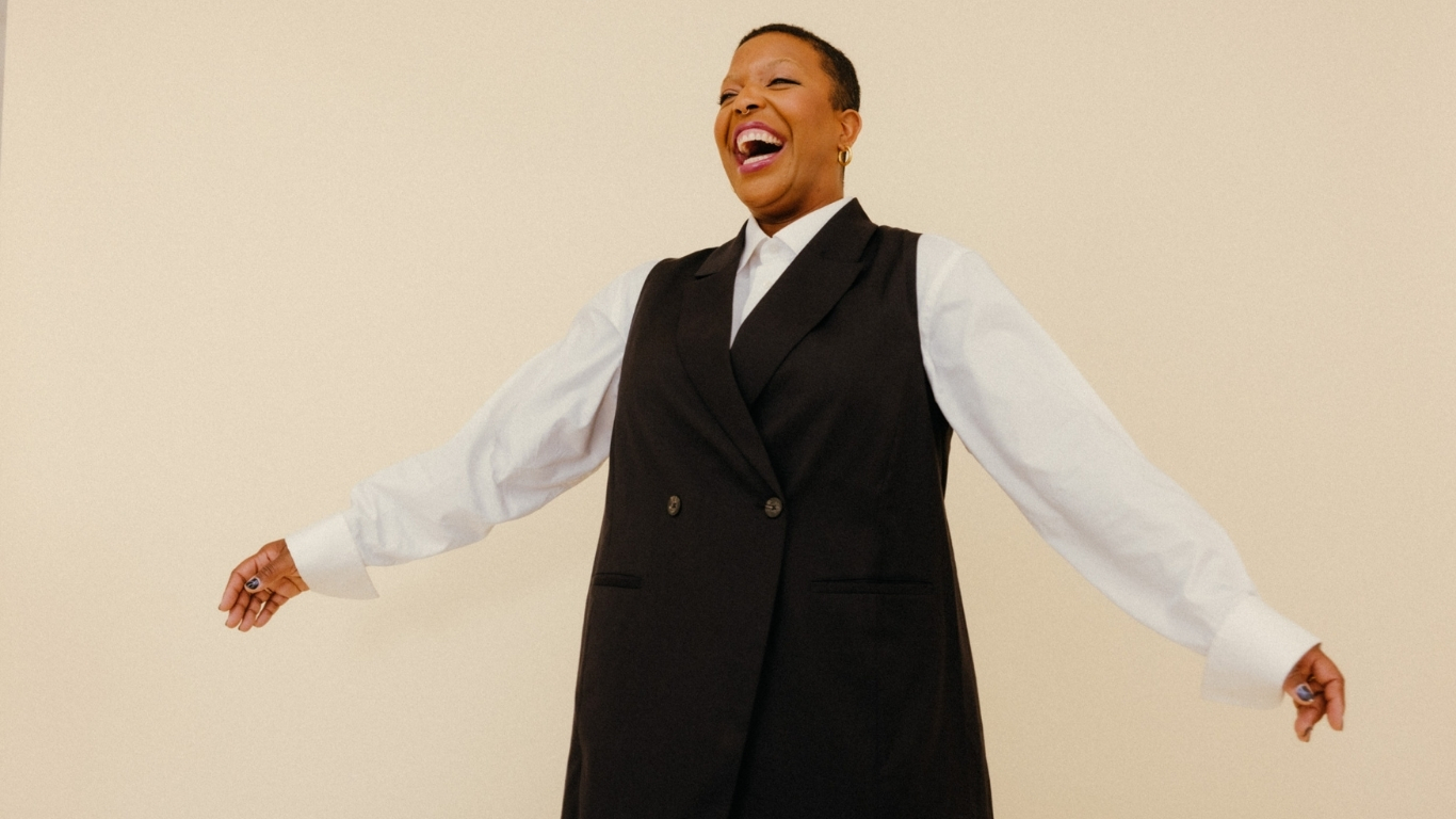 A woman with short hair, wearing a white shirt and a black vest, is smiling and extending her arms outwards against a beige background.