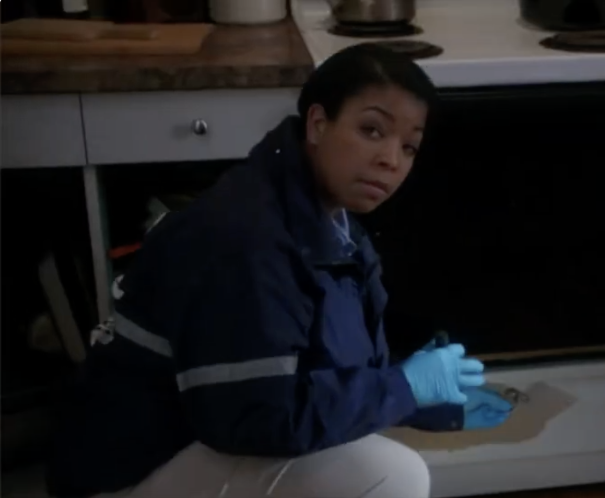 A woman with short dark hair wearing a dark jacket and blue gloves is kneeling beside a kitchen island, looking at the camera with a serious expression.