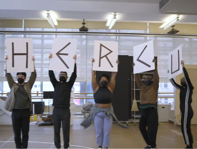 Group of five dancers holding large white signs with black letters spelling 'H E R E L U' in a dance studio.