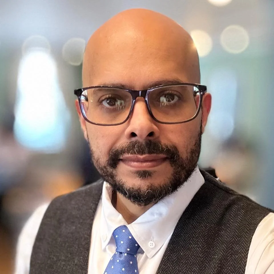 A man with glasses, a bald head, and a beard, dressed in a white shirt and blue polka-dot tie, poses for a selfie in a setting with blurred lights in the background.