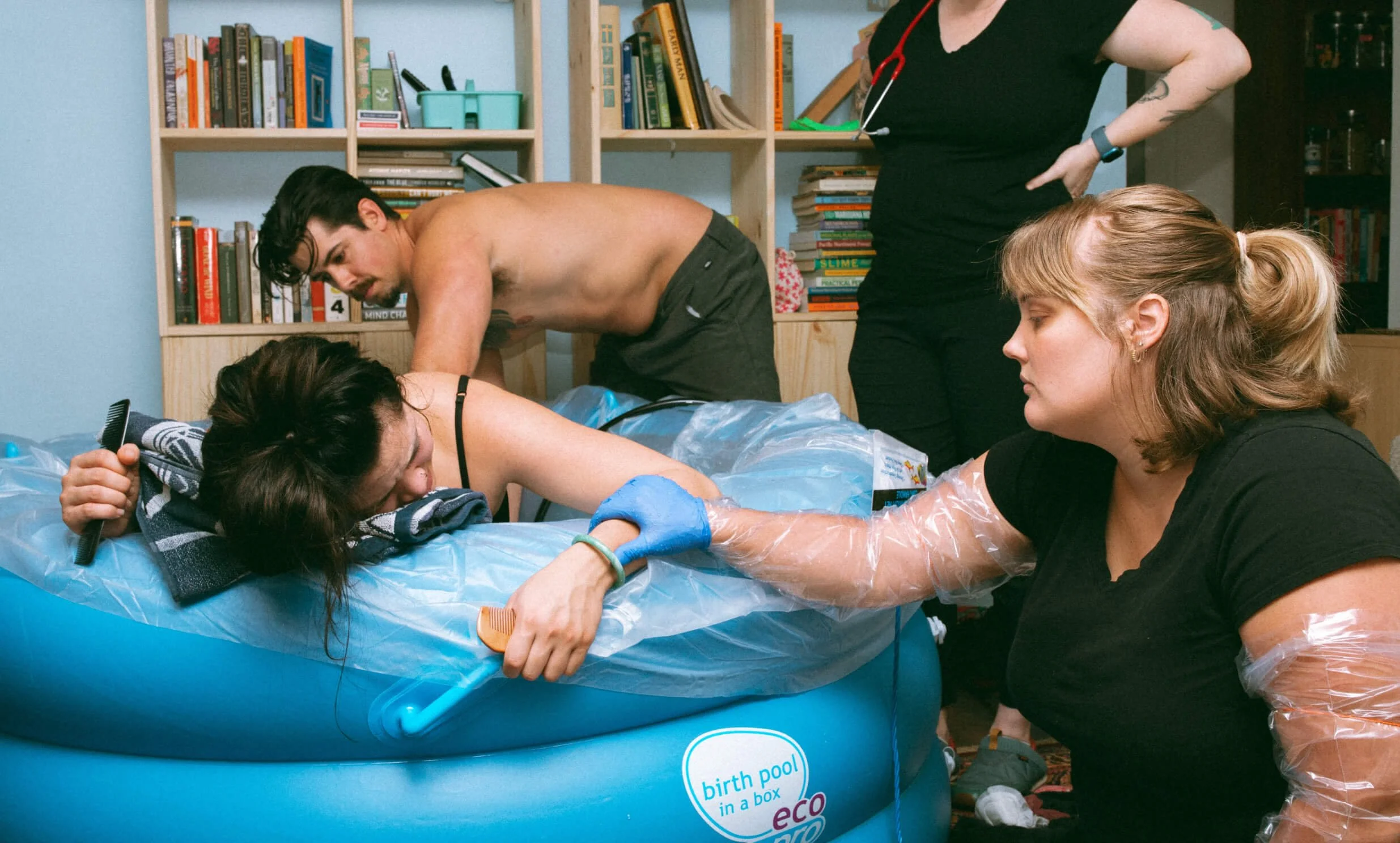 A woman in a birthing pool is supported by a partner and a birth assistant, who is wearing gloves and plastic sleeves, during a home water birth.