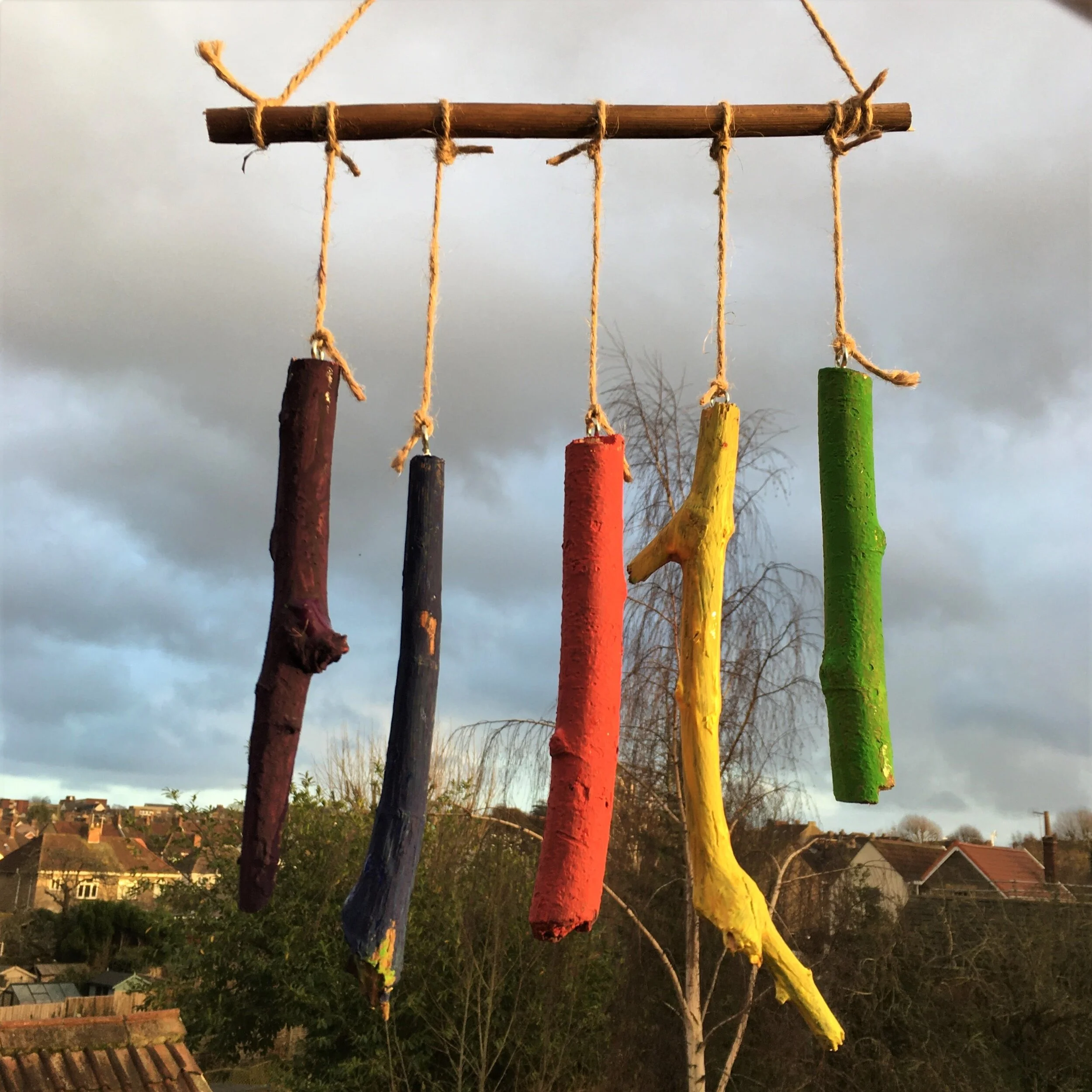 Colorful painted sticks hanging from a wooden rod with twine, set against a cloudy sky and residential neighborhood in the background.