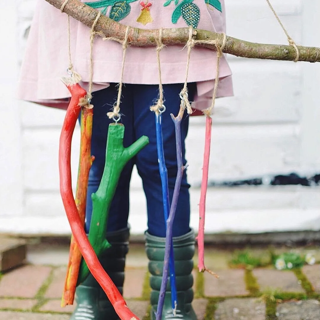 Child wearing rubber boots and dark pants, holding a stick with colorful painted sticks hanging from it, outdoors against a white fence.