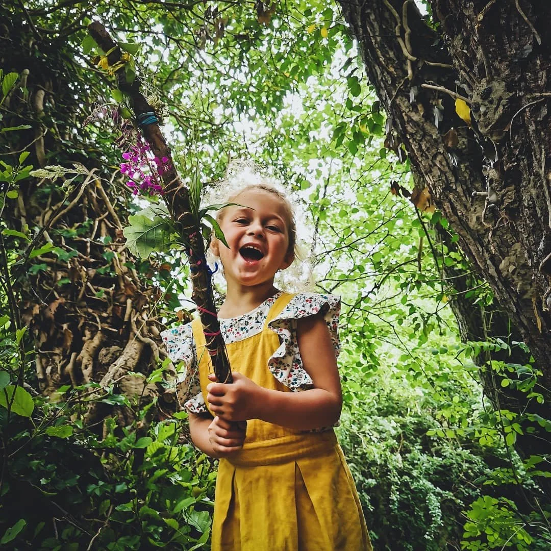 A young girl with curly blonde hair smiling and holding a decorated stick, standing amidst dense green foliage in a forest.